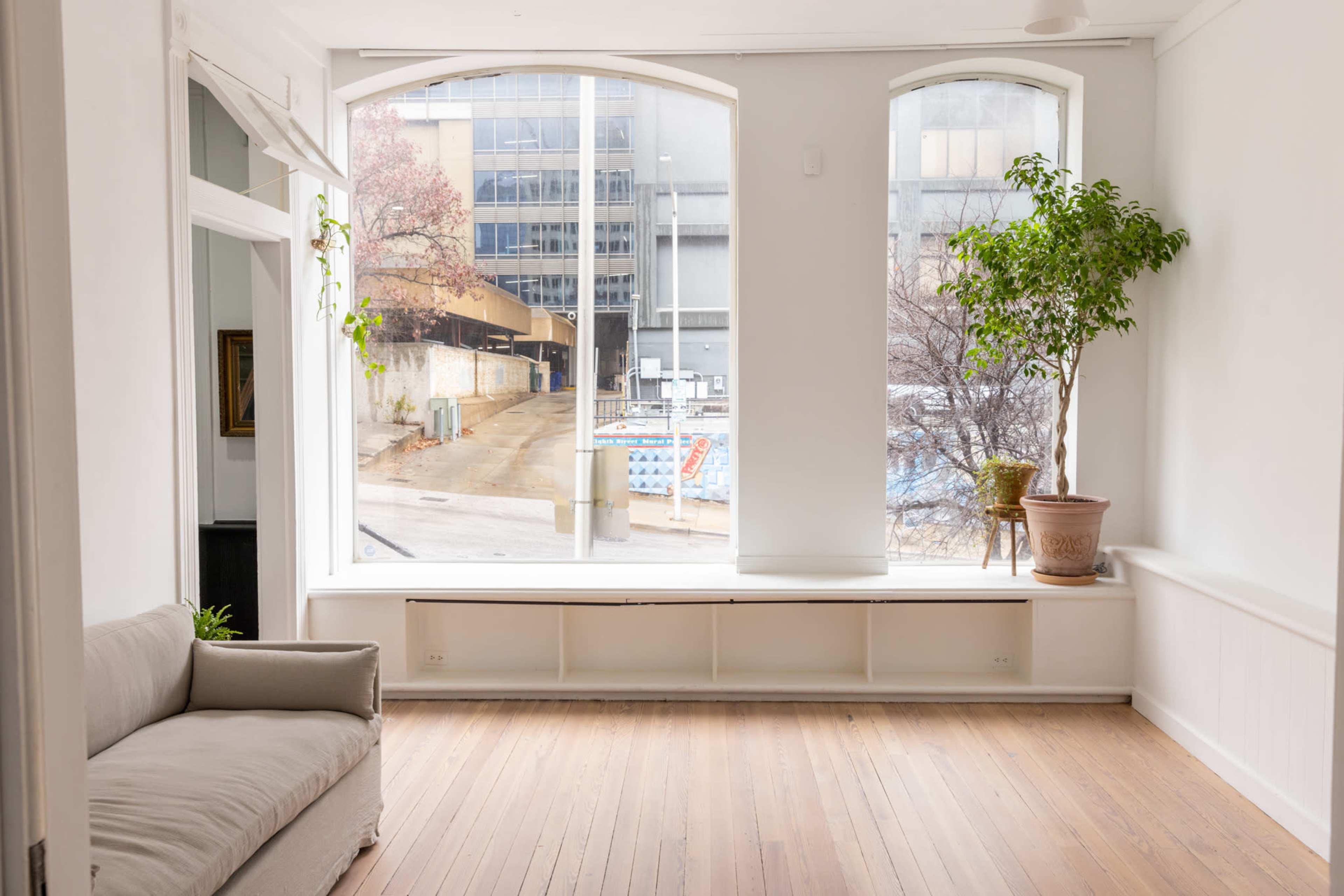 A bright, minimalist interior with large windows overlooking a street and a potted plant beside a light-colored sofa.