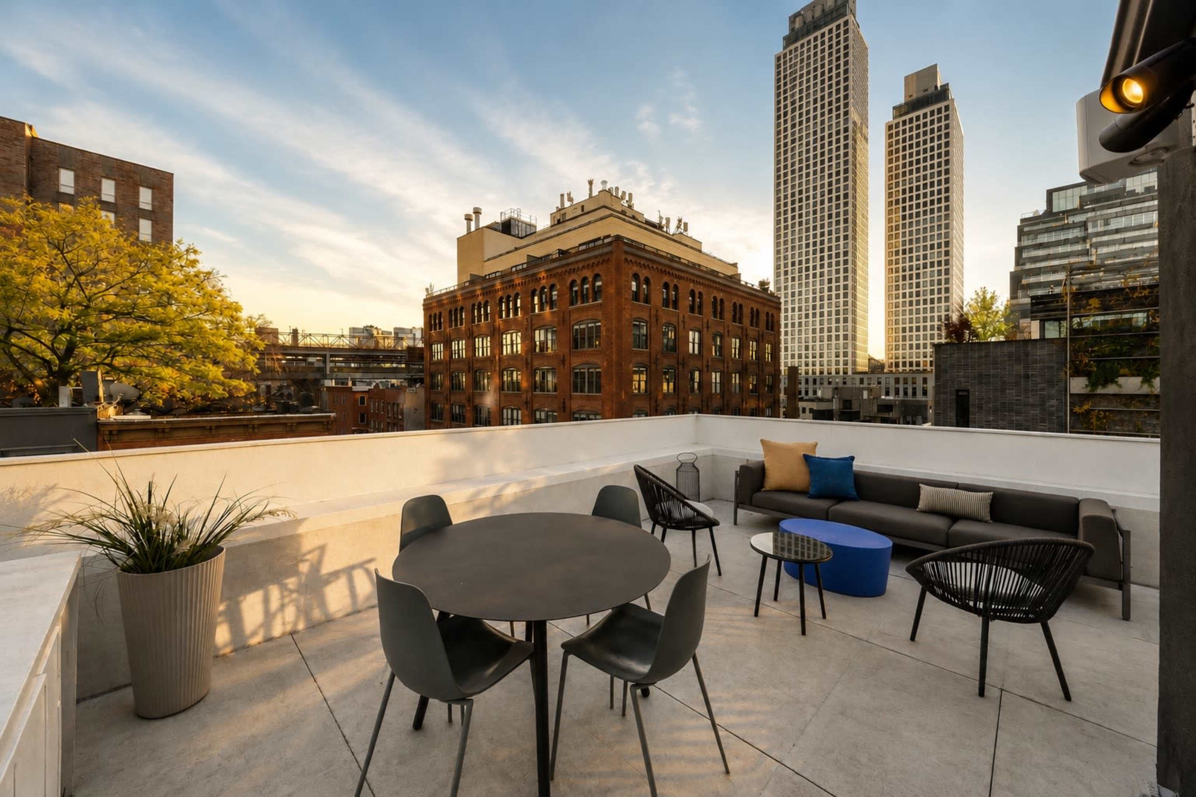 A rooftop terrace features a circular table surrounded by chairs, with a view of historical and modern buildings against a sunset sky.