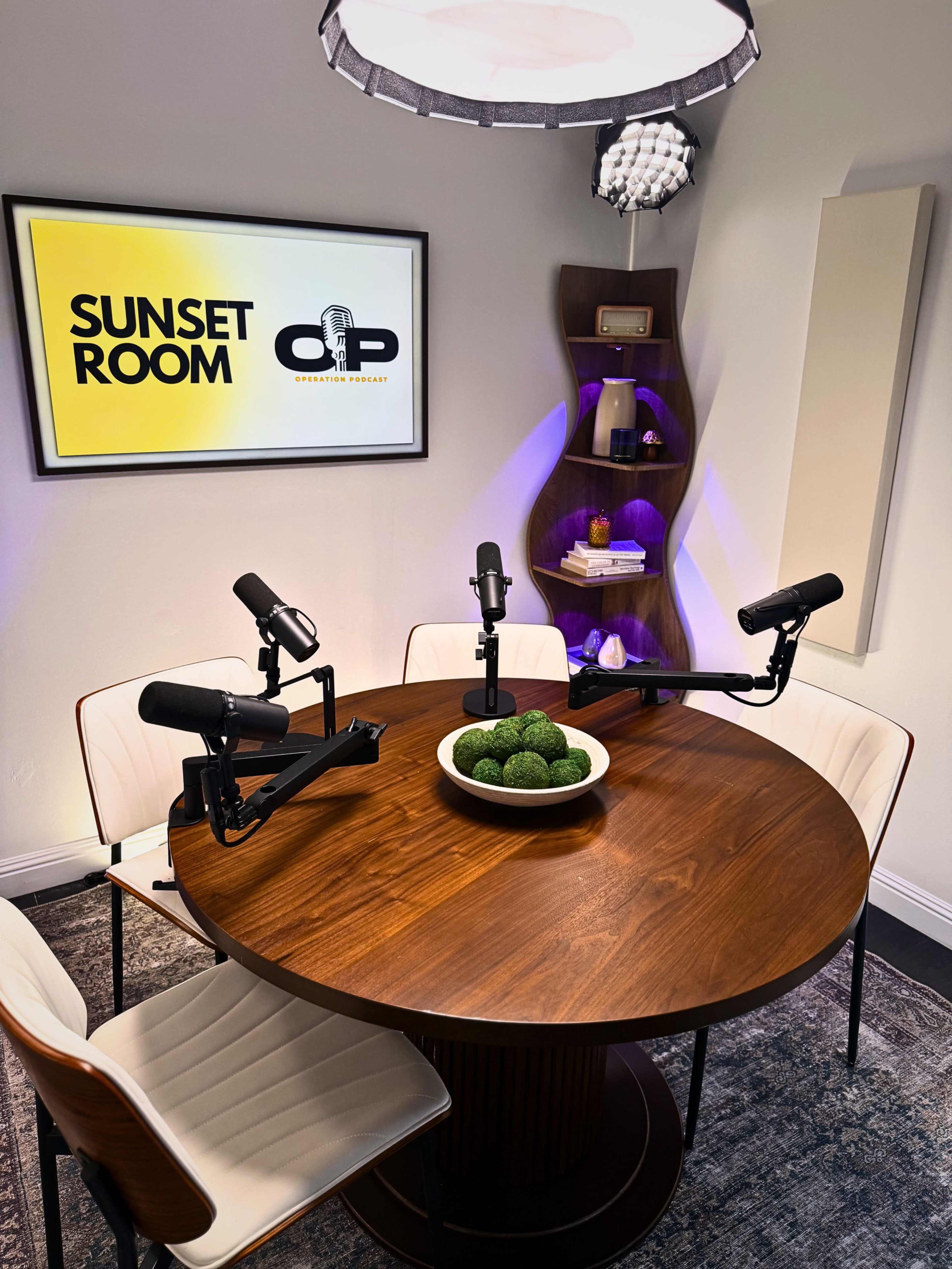 The image shows a modern podcast recording studio featuring a round wooden table with four microphones positioned around it, a decorative bowl of greenery in the center, and a wall-mounted display labeled "SUNSET ROOM."