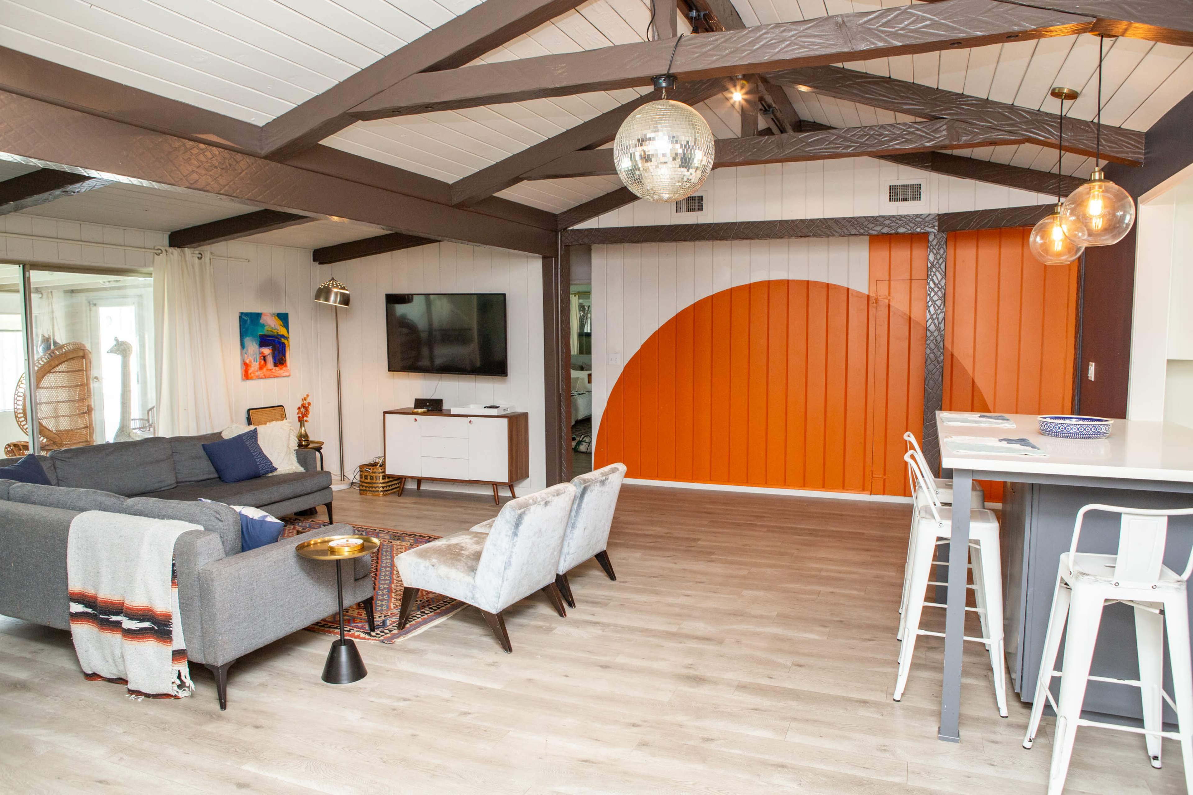 A modern living area featuring a gray sofa, a large flat-screen TV, and an orange accent wall, with a dining area that includes white bar stools.