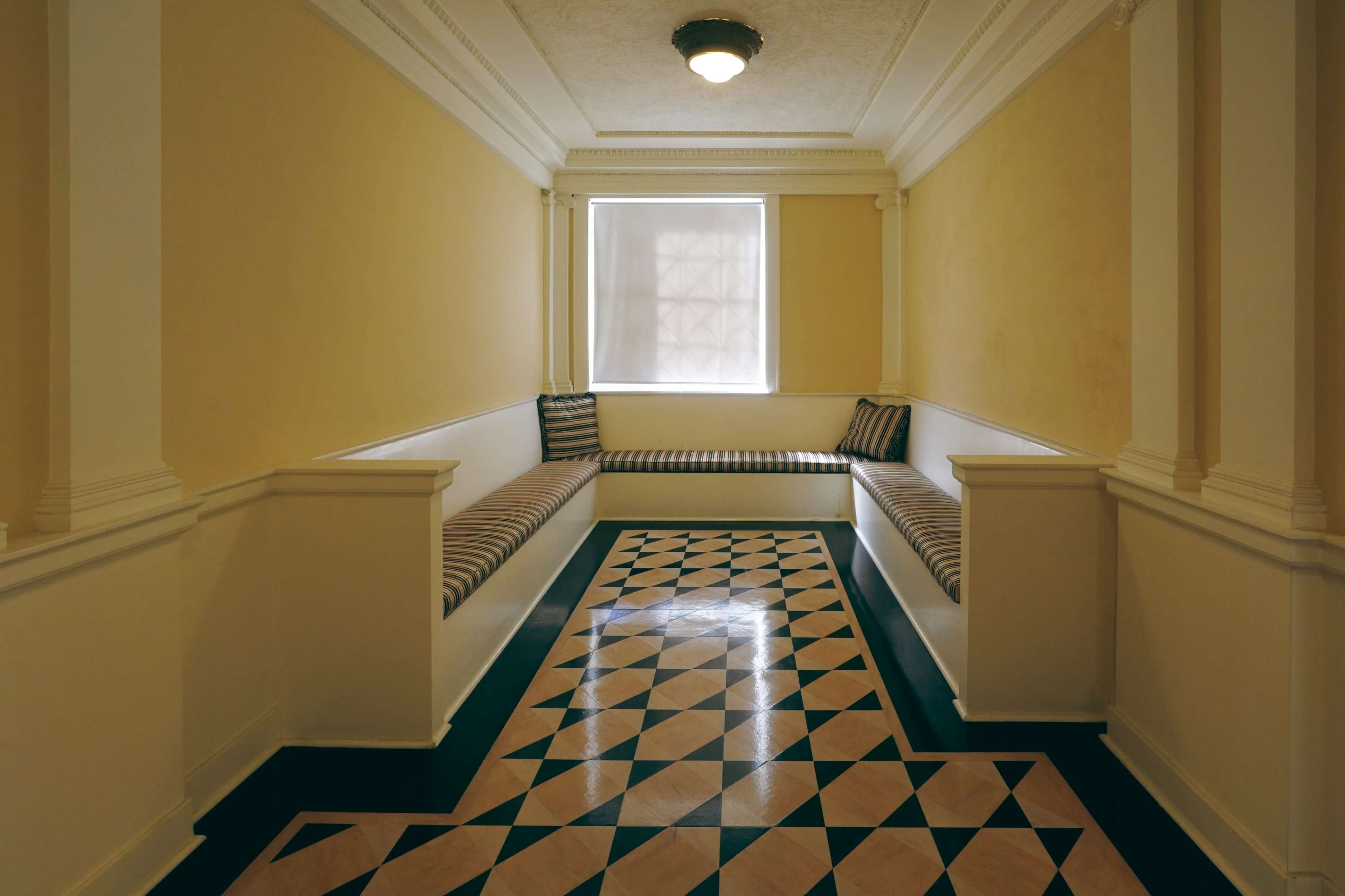 A narrow corridor with geometric patterned flooring and built-in seating along the walls under a window.