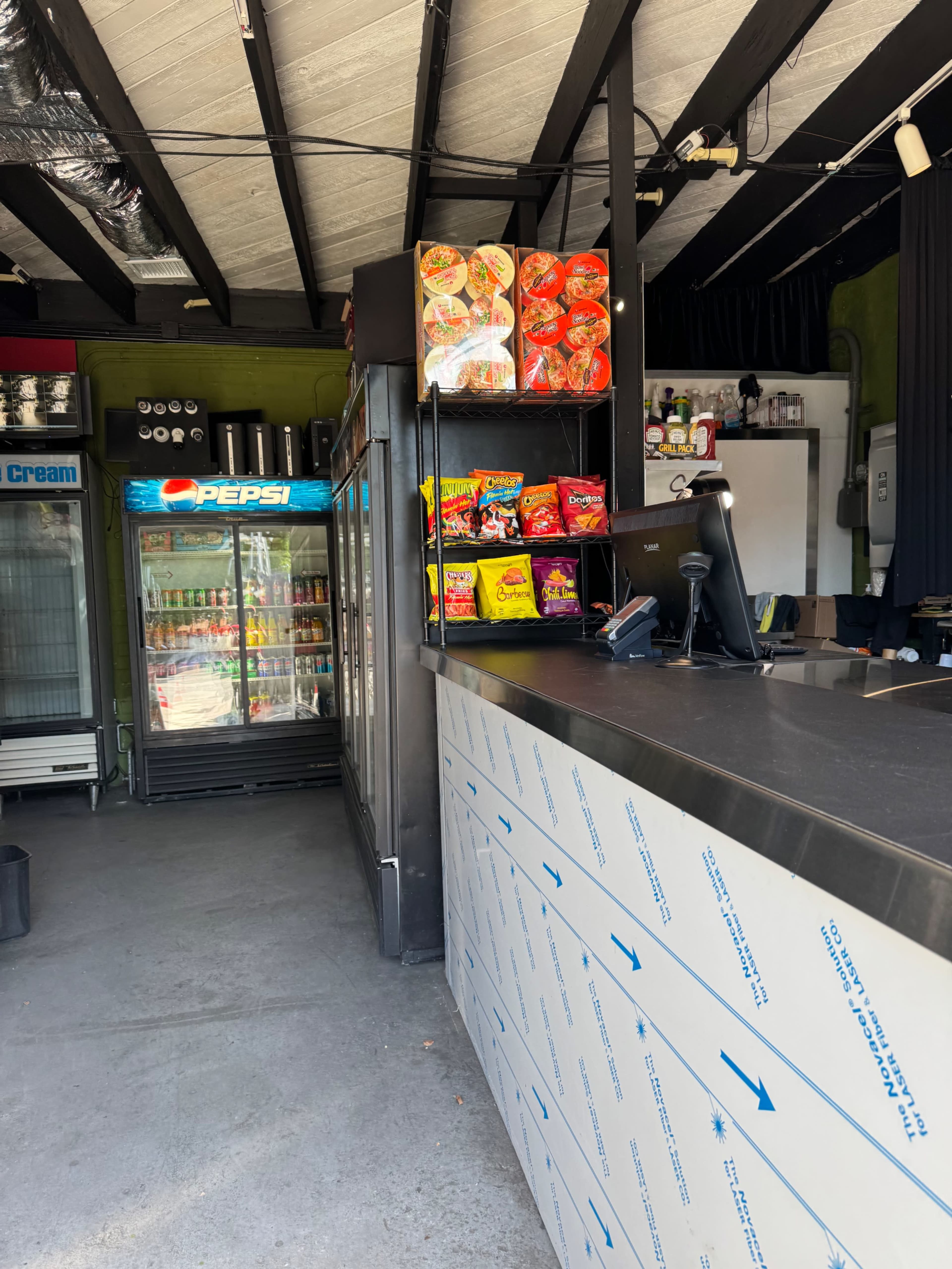 The image shows the interior of a small convenience store with a service counter, various snacks on display, and a beverage cooler featuring Pepsi products.