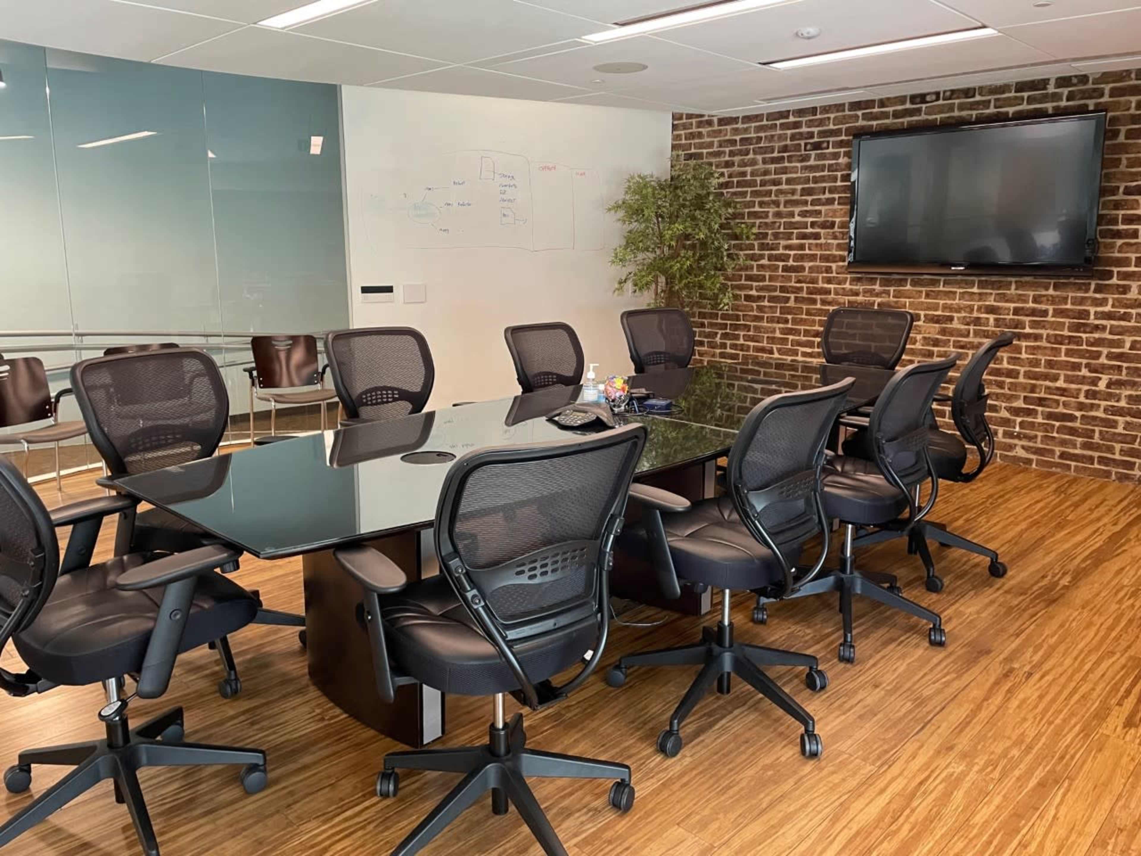 A modern conference room features a large glass table surrounded by ergonomic chairs, with a brick wall and a mounted television.