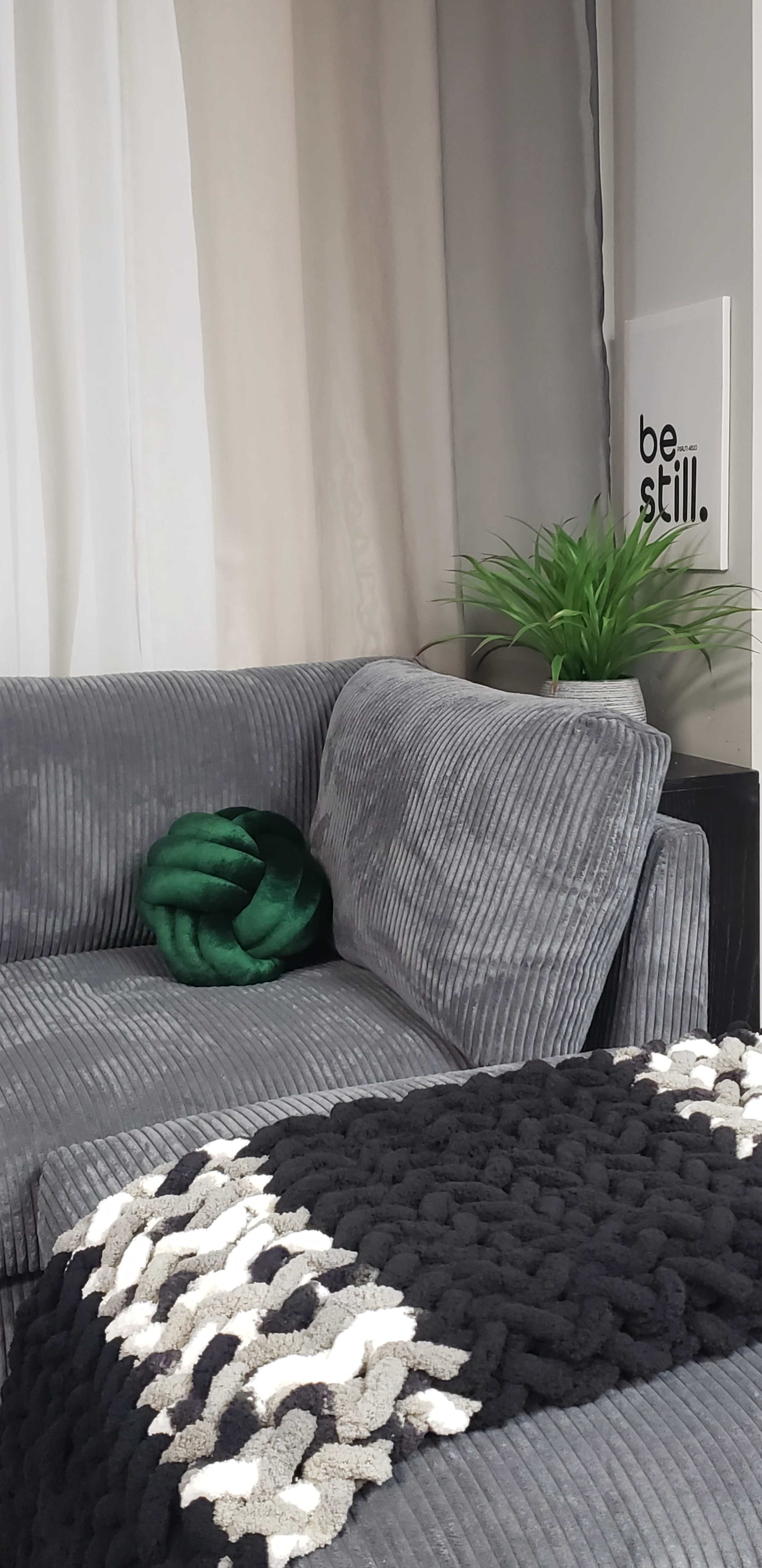 A gray corduroy couch with a green knotted pillow and a black and white textured throw blanket sits beside a potted plant and a wall with the text "be still."
