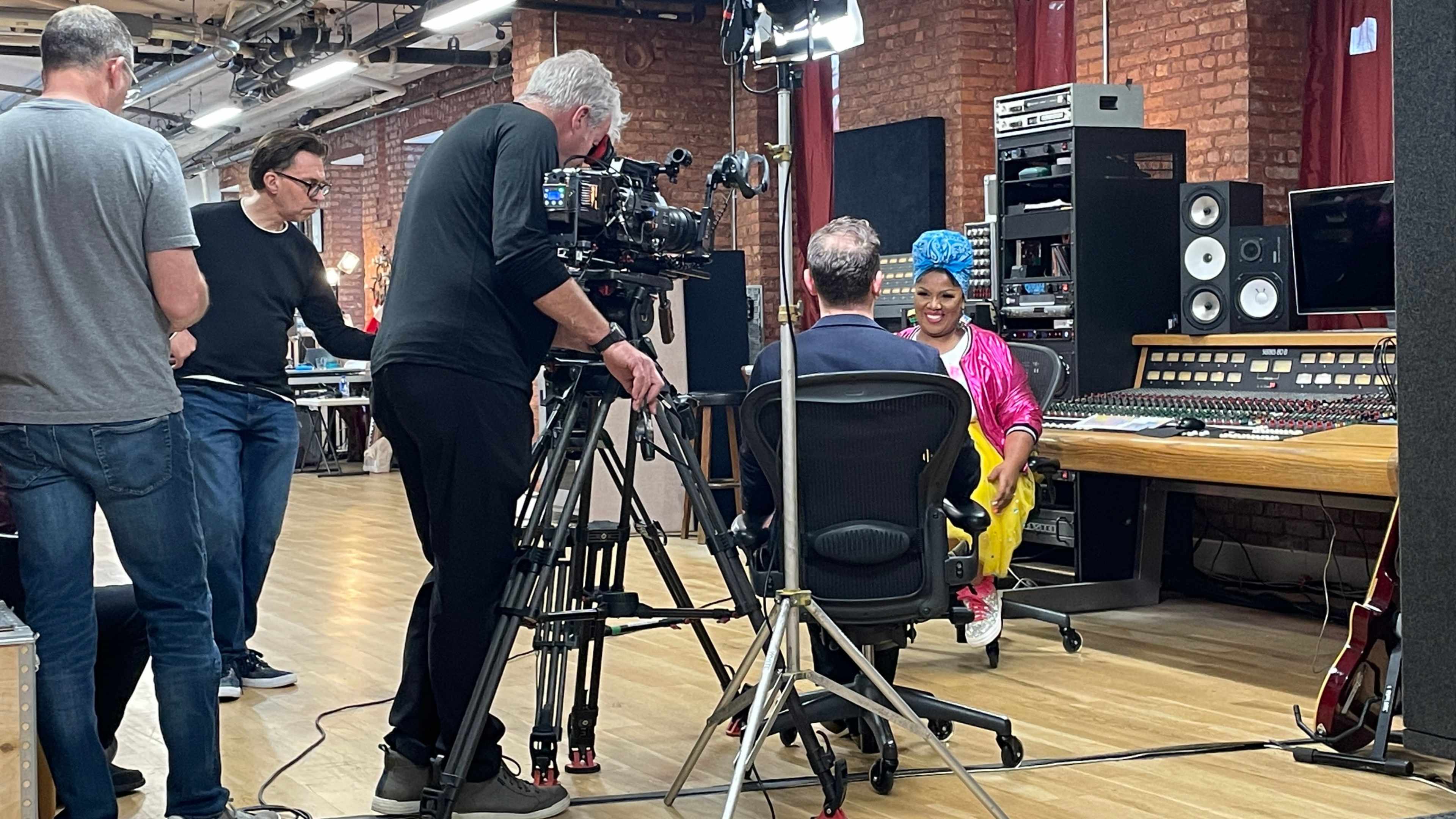 A film crew records an interview in a studio filled with audio equipment and musical instruments.