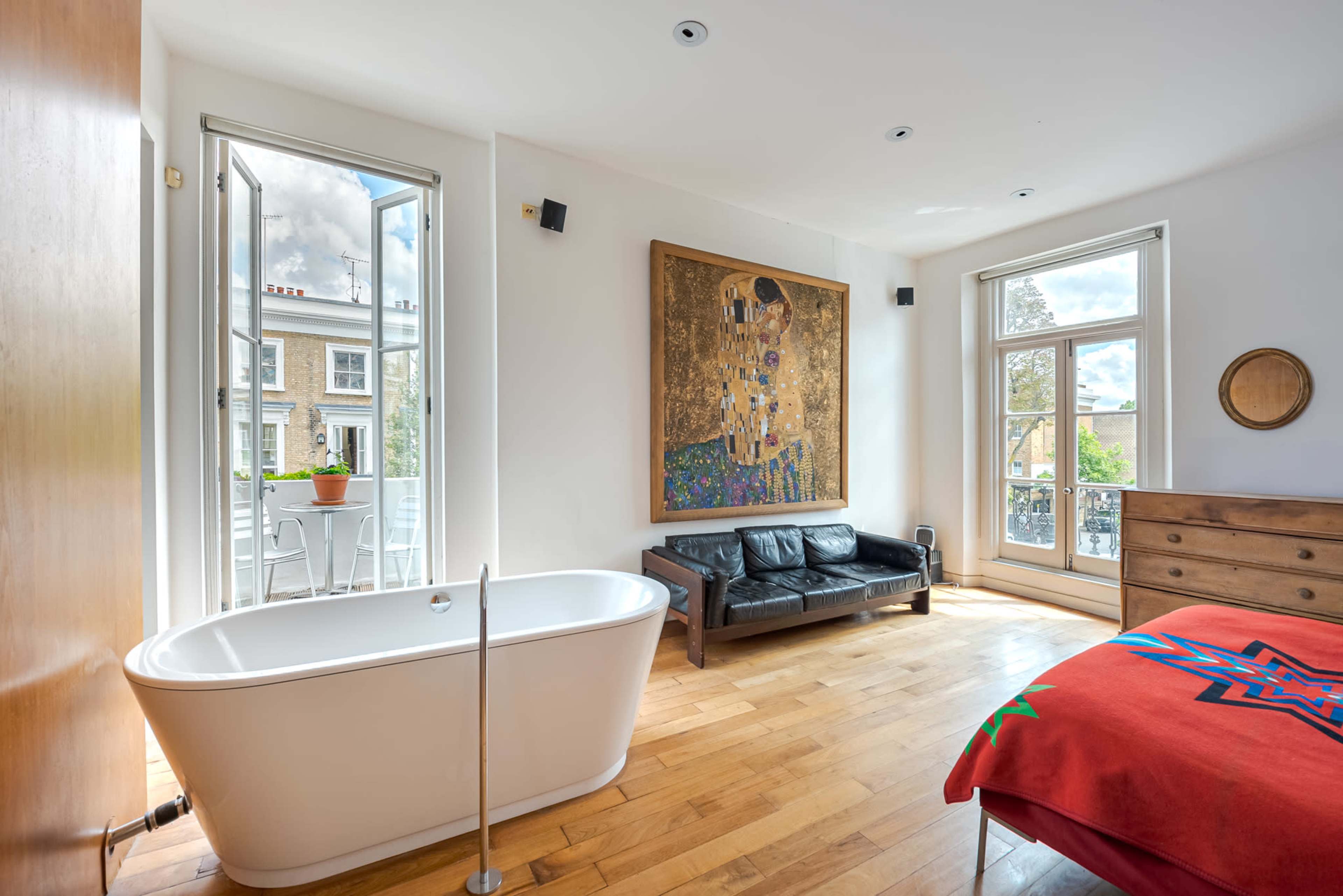 A stylish bedroom features a freestanding bathtub, a black leather sofa, and large windows with a view of a balcony, complemented by a prominent piece of artwork on the wall.