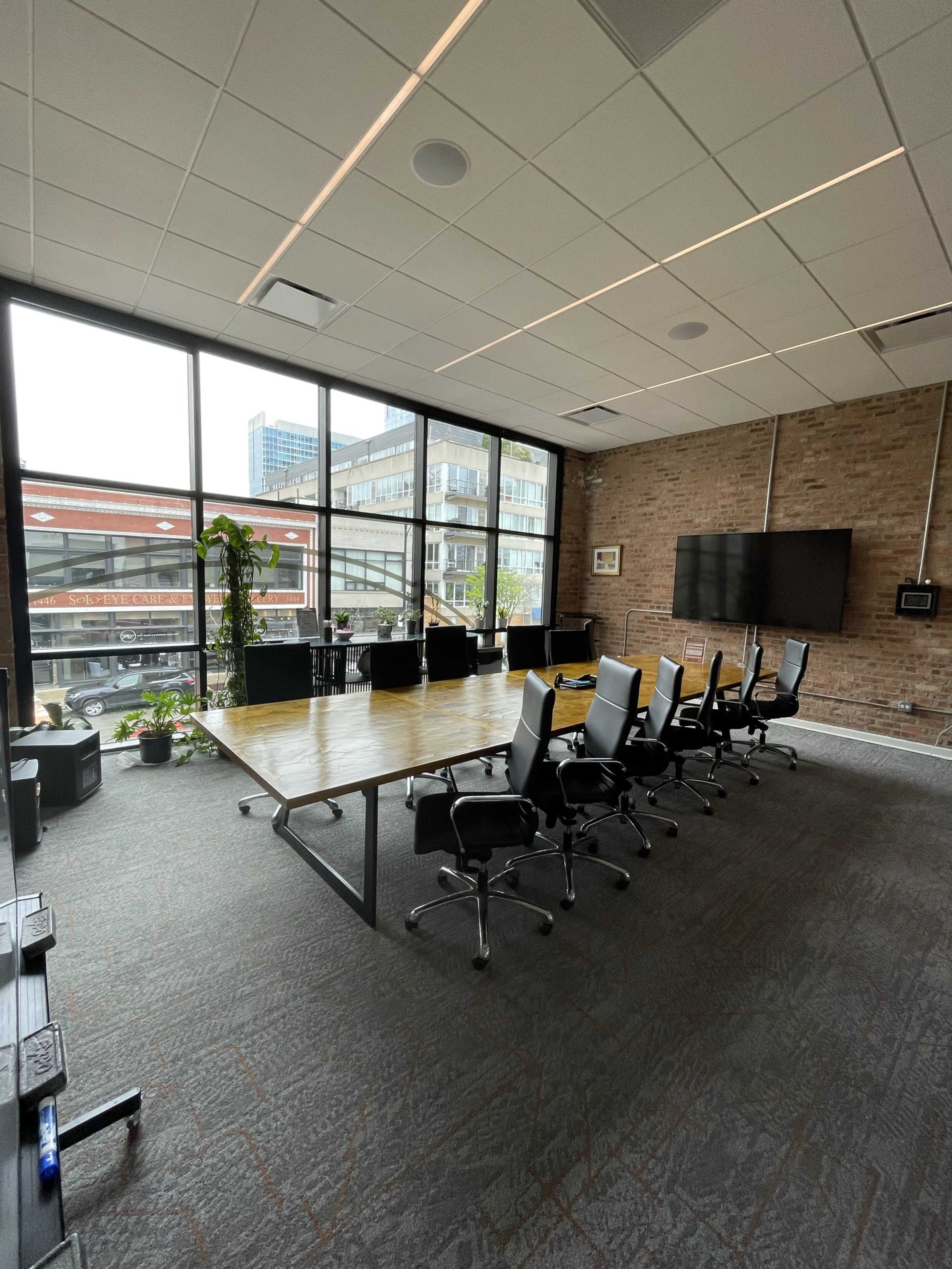 A modern conference room features a large wooden table surrounded by black office chairs, with floor-to-ceiling windows allowing natural light and a view of a city landscape.