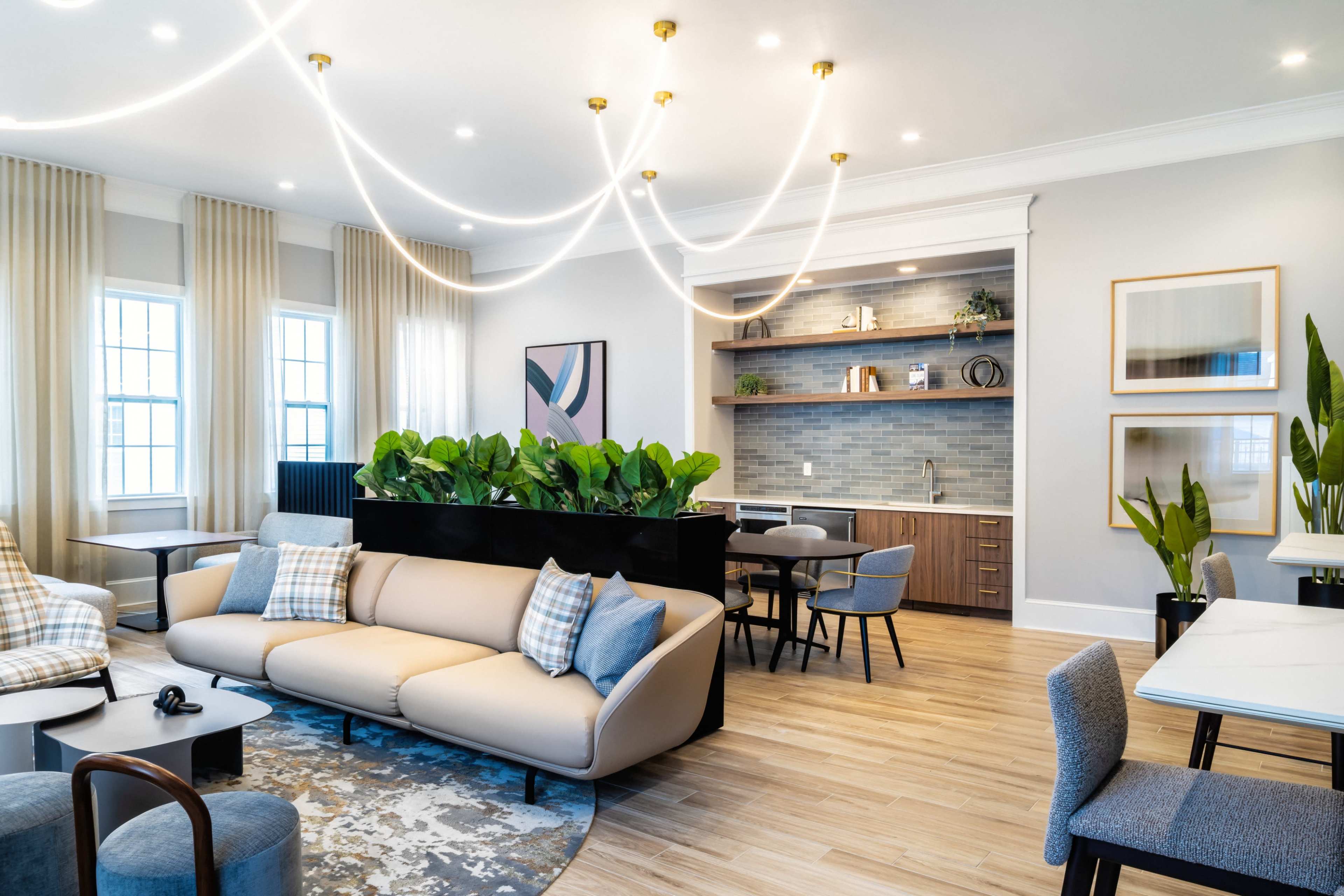 The image shows a modern living space featuring a light-colored sofa, several plants, a kitchen area with shelves, and various seating arrangements.