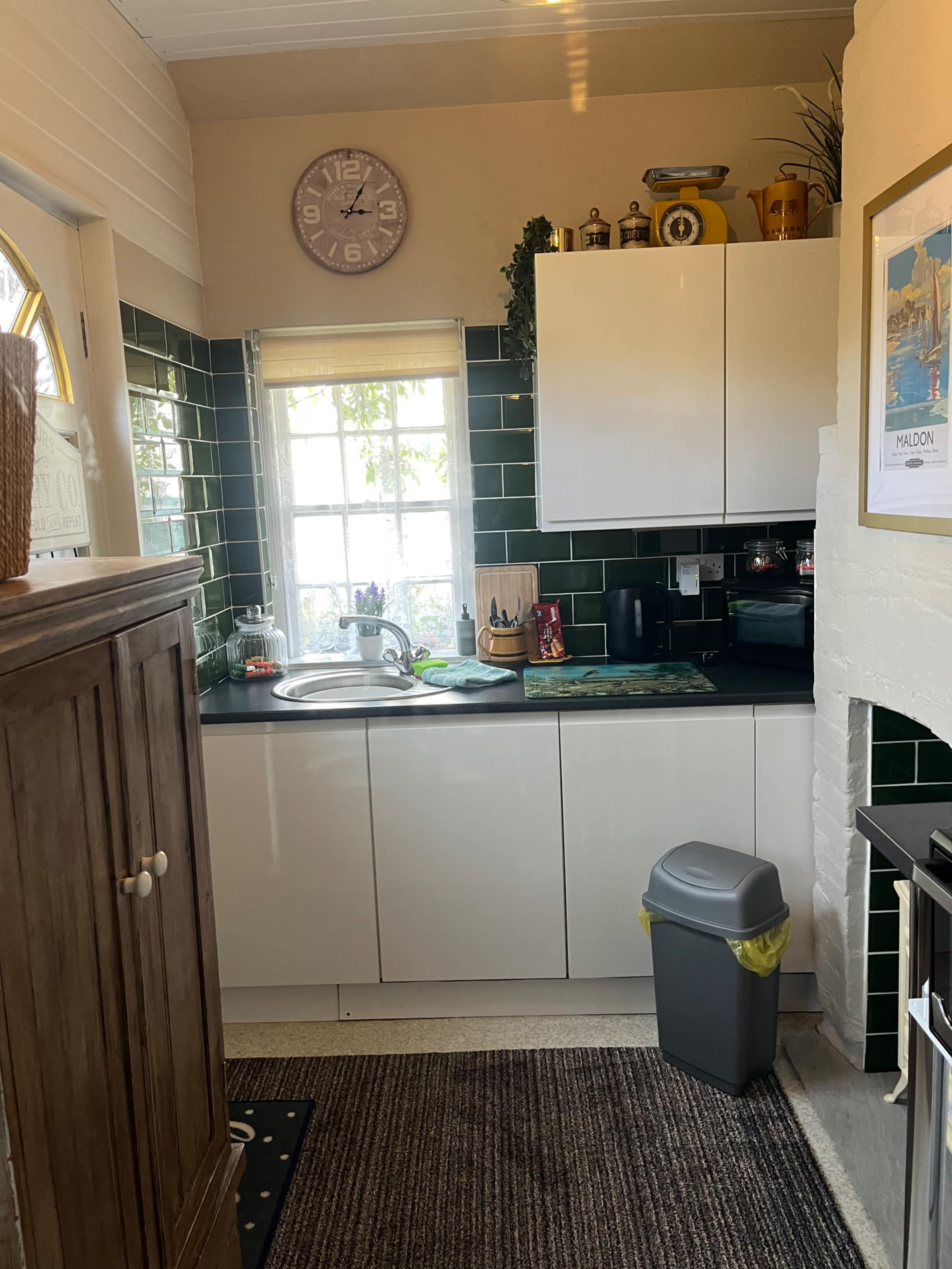 The image shows a compact kitchen with white cabinetry, green tiled walls, and a stainless steel sink, featuring a small window and a wooden storage cabinet.