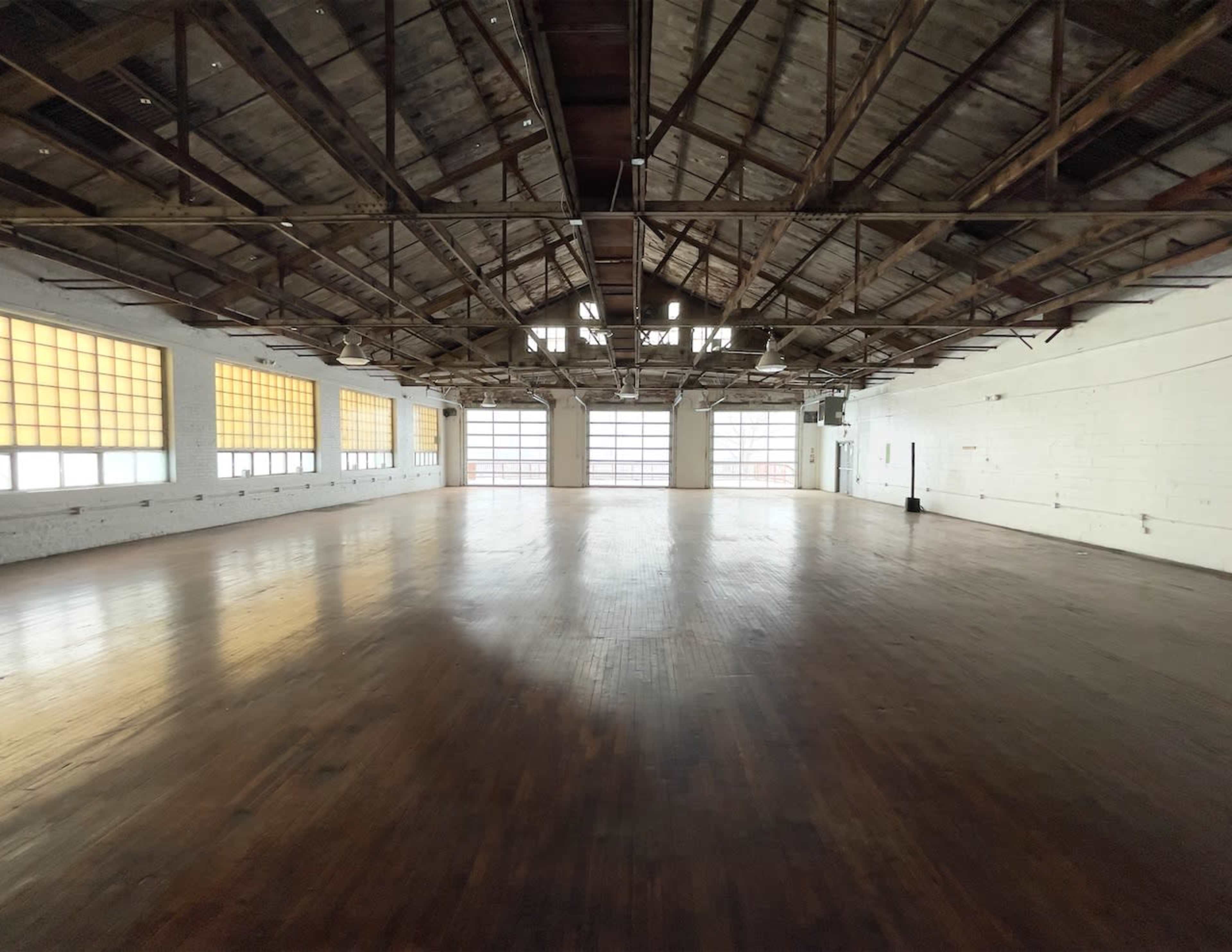 A spacious, empty room with wooden floors and industrial-style windows along the walls, featuring exposed beams and a high ceiling.