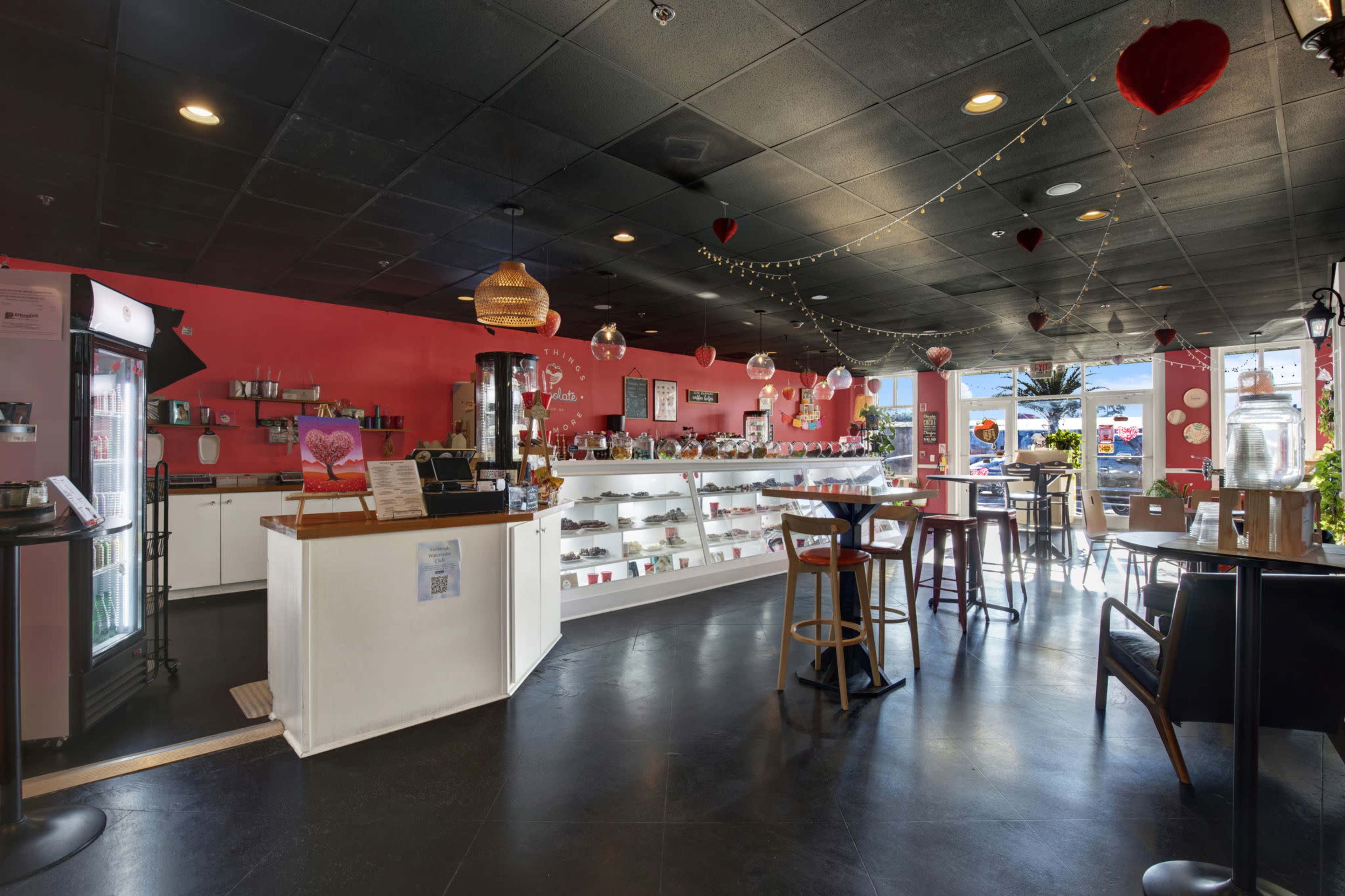 The image shows a brightly colored cafe with a counter displaying various desserts and drinks, decorated with heart-shaped ornaments and large windows allowing natural light to enter.