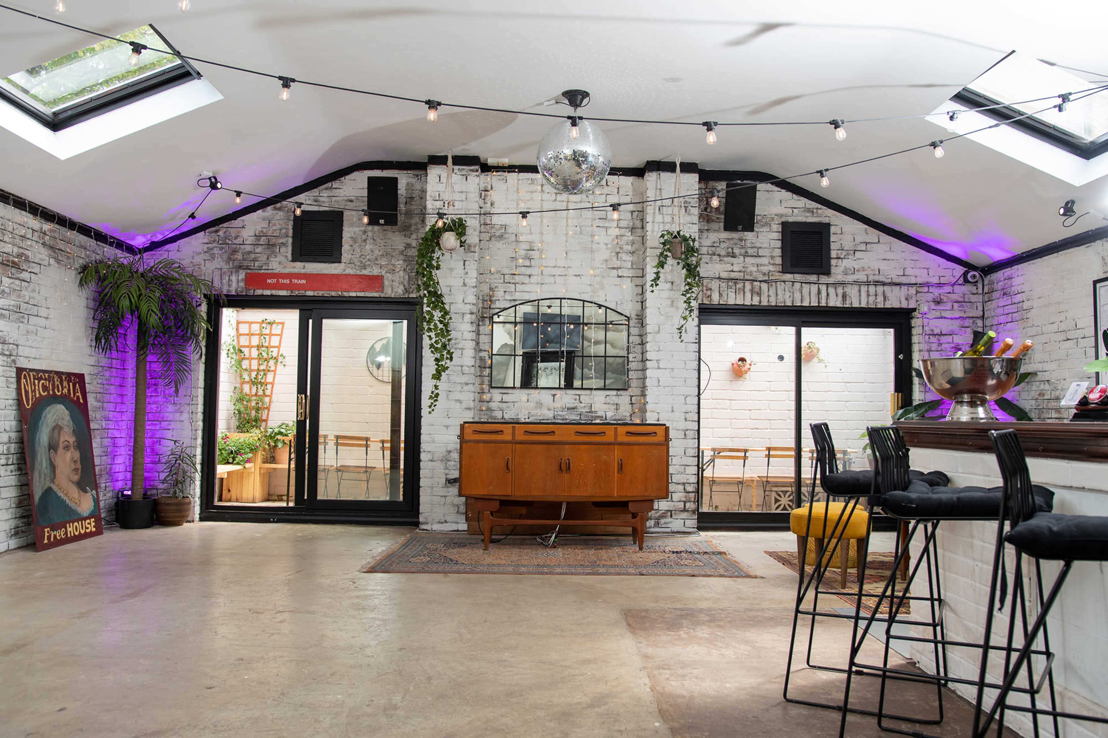 A spacious, modern interior with a bar area, large windows, and decorative lighting, featuring a mix of white brick walls and greenery.