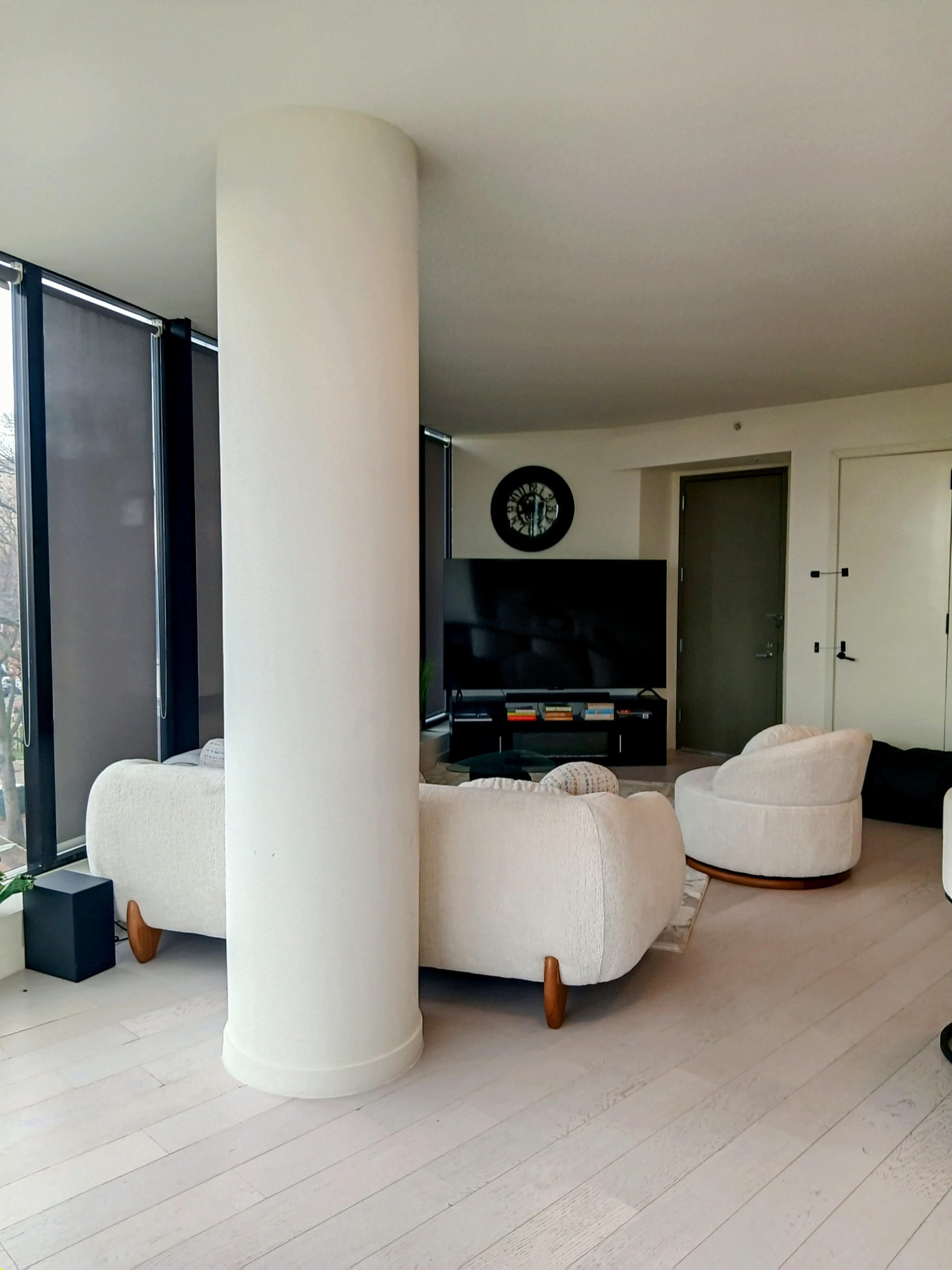A cozy living area features two white, rounded chairs, a television on a low console, and large windows with light-filtering shades.
