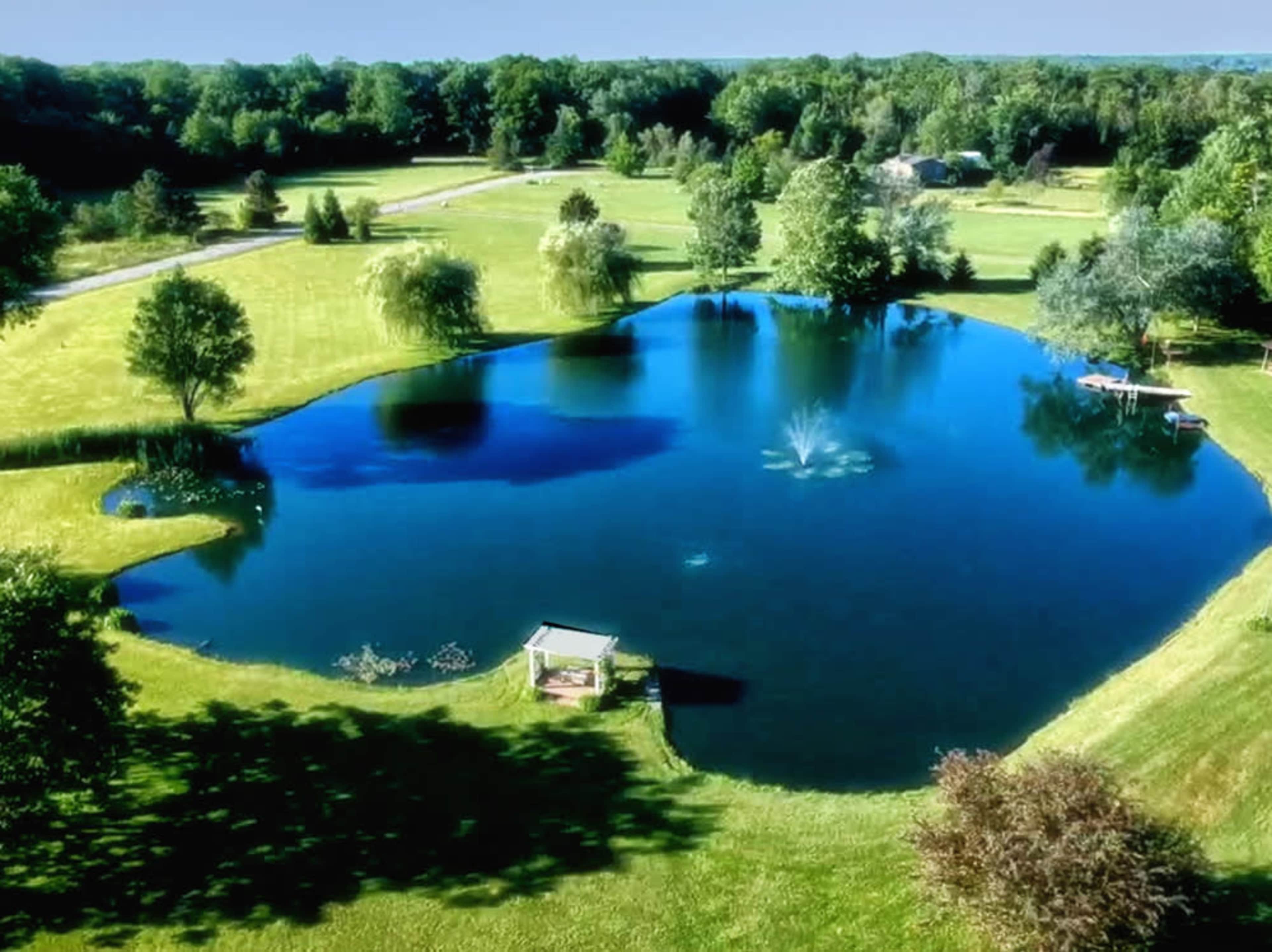 A large pond surrounded by grass and trees, with a fountain in the center and a gazebo nearby.