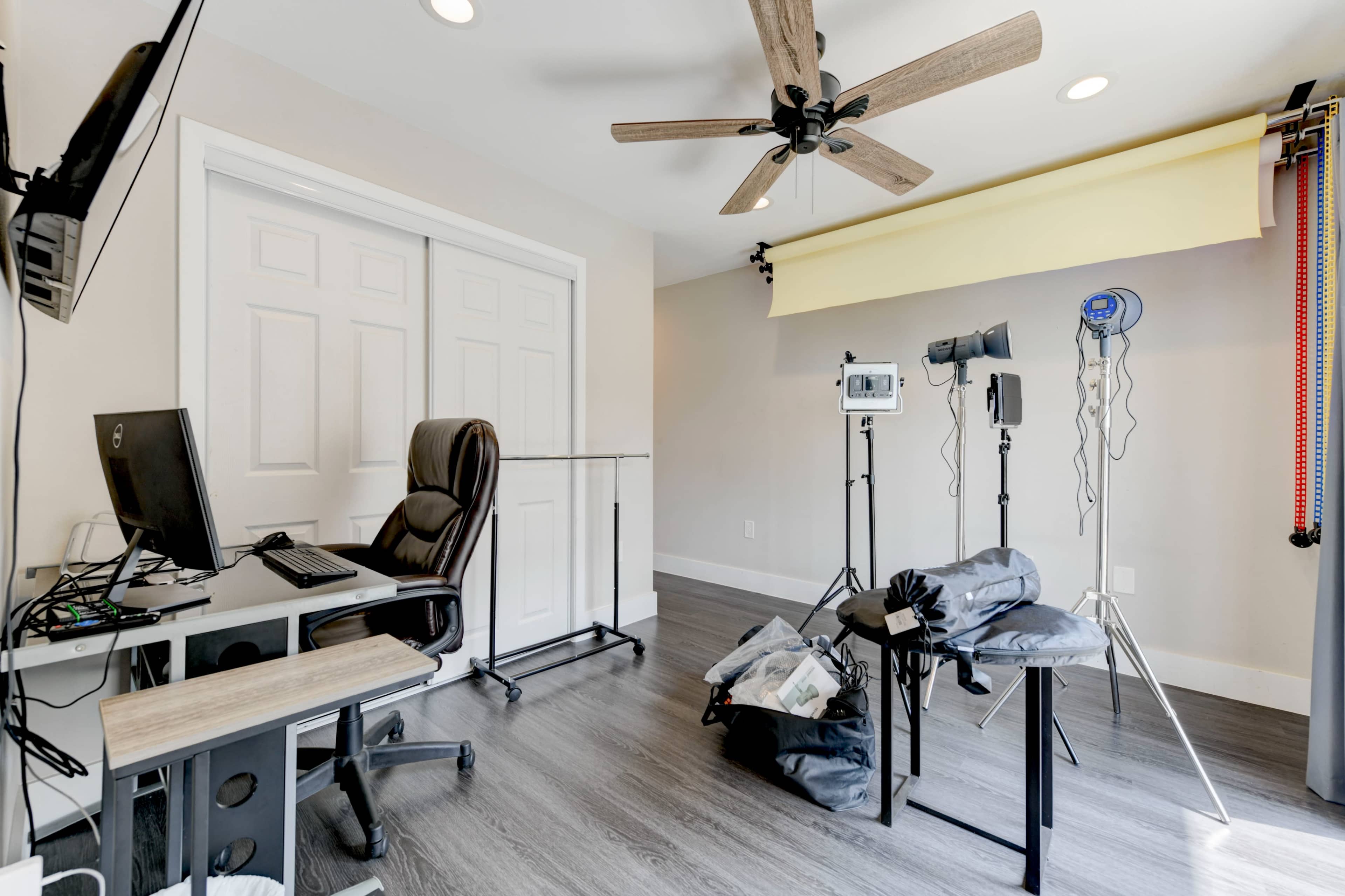 A workspace featuring a desk with a computer and chair, a clothing rack, and photography equipment including lights and a backdrop.