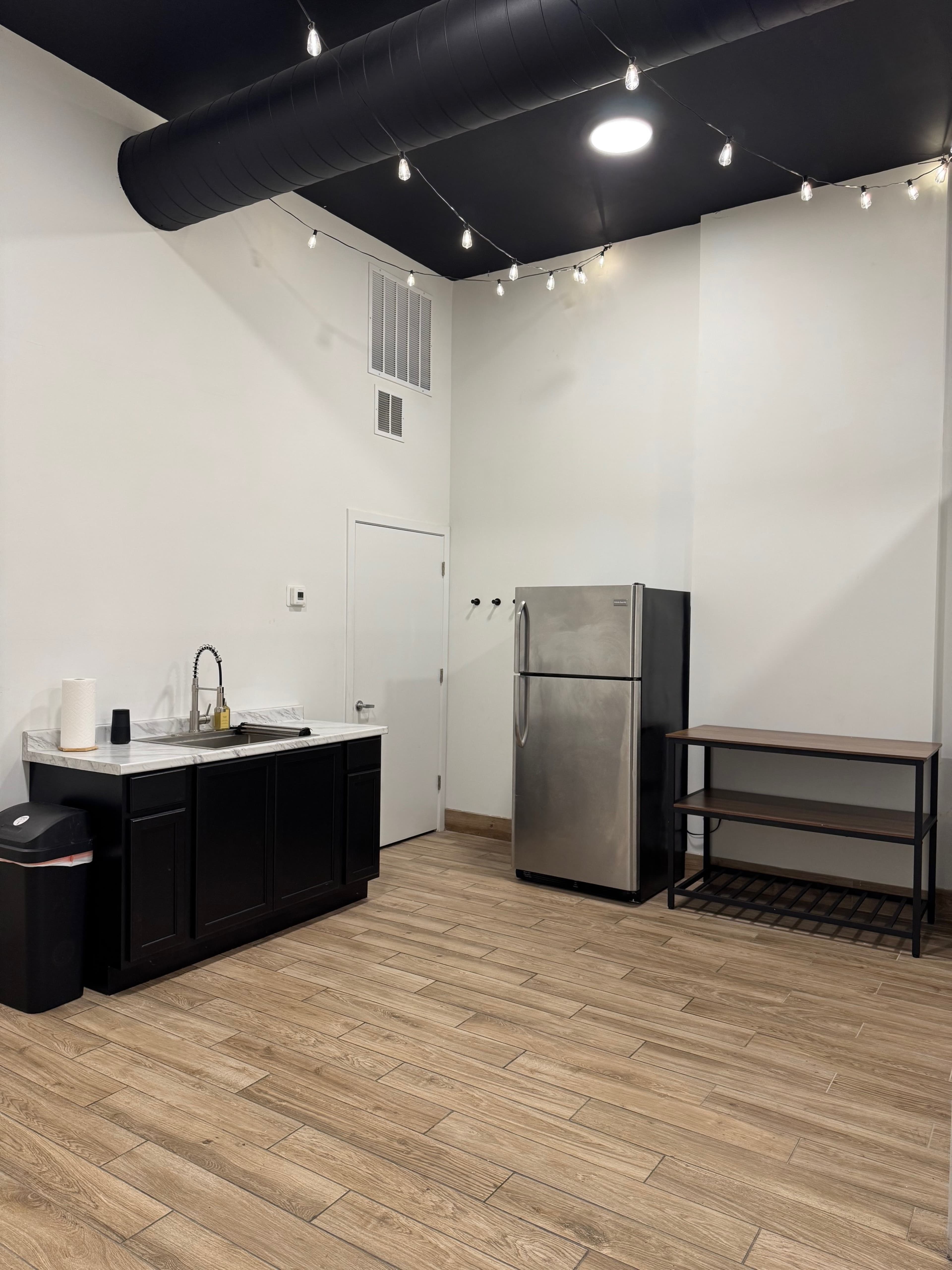 The image shows a kitchen area with a silver refrigerator, a dark cabinet under a sink, a black trash can, and a metal shelf, all set against a white wall and a ceiling with exposed black ductwork and string lights.