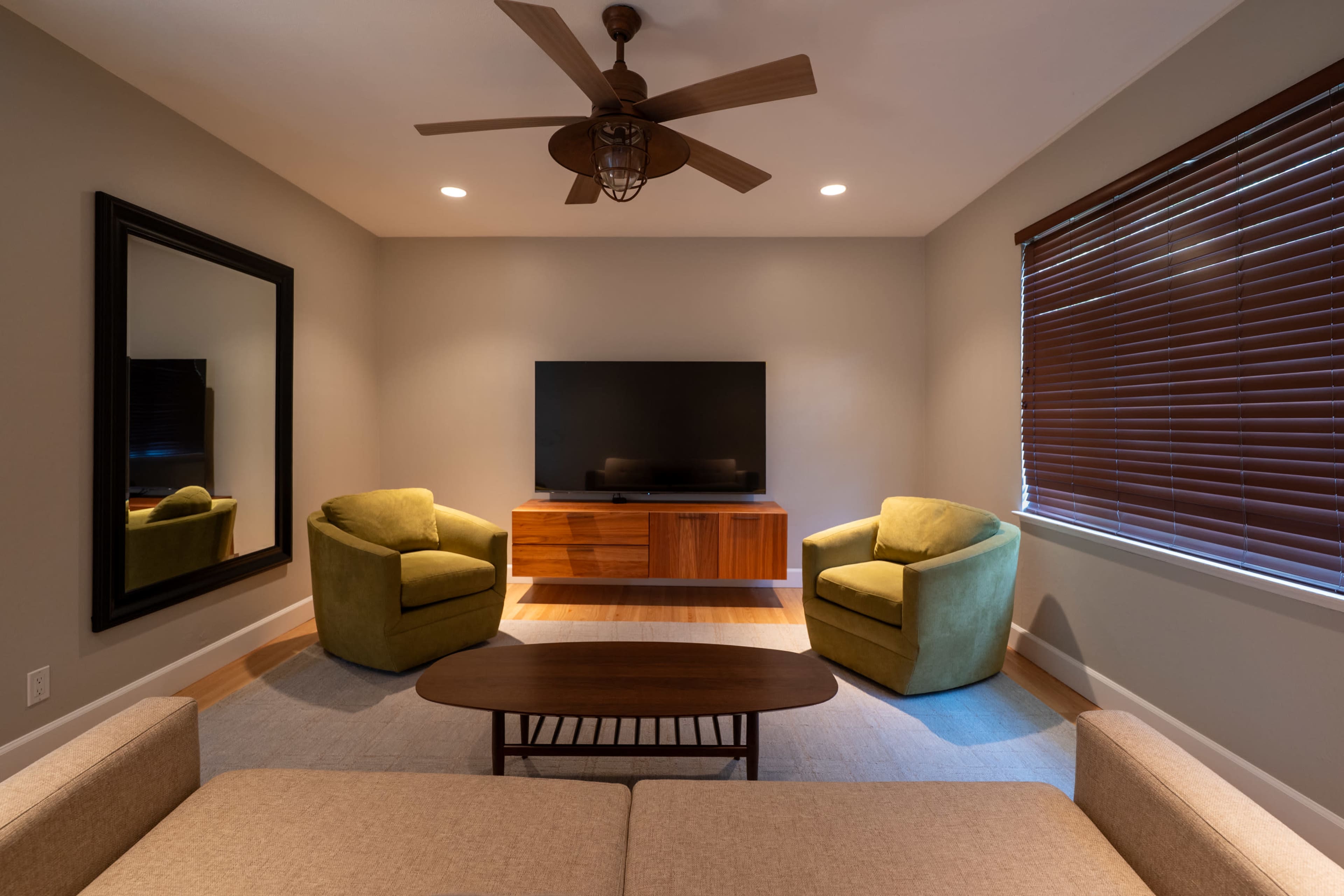 The image shows a modern living room featuring two green armchairs, a brown coffee table, a flat-screen TV mounted on the wall, and a large mirror.