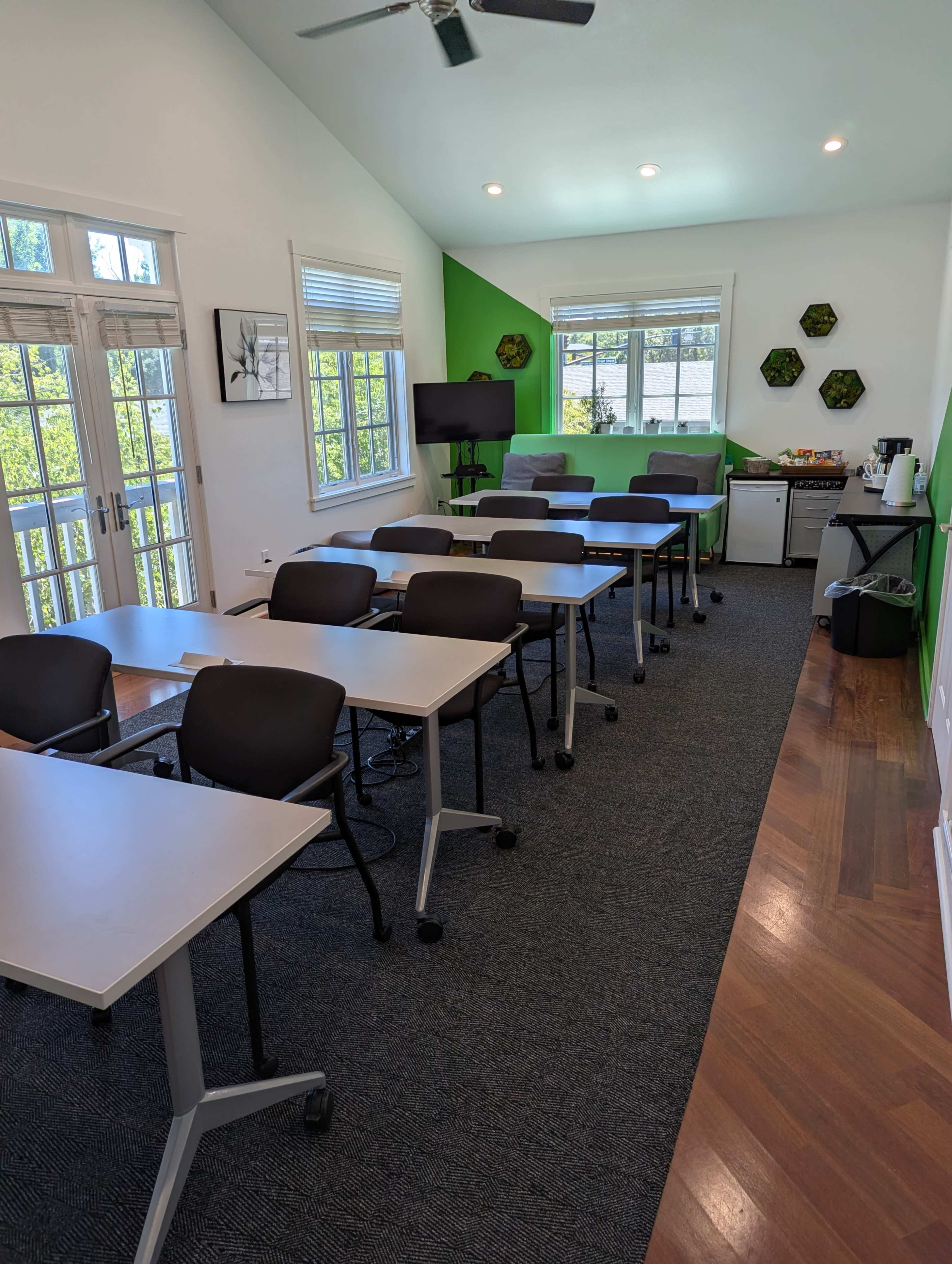 The image shows a bright classroom or meeting space with several tables and chairs arranged in rows, a small kitchenette area, and large windows allowing natural light to enter.