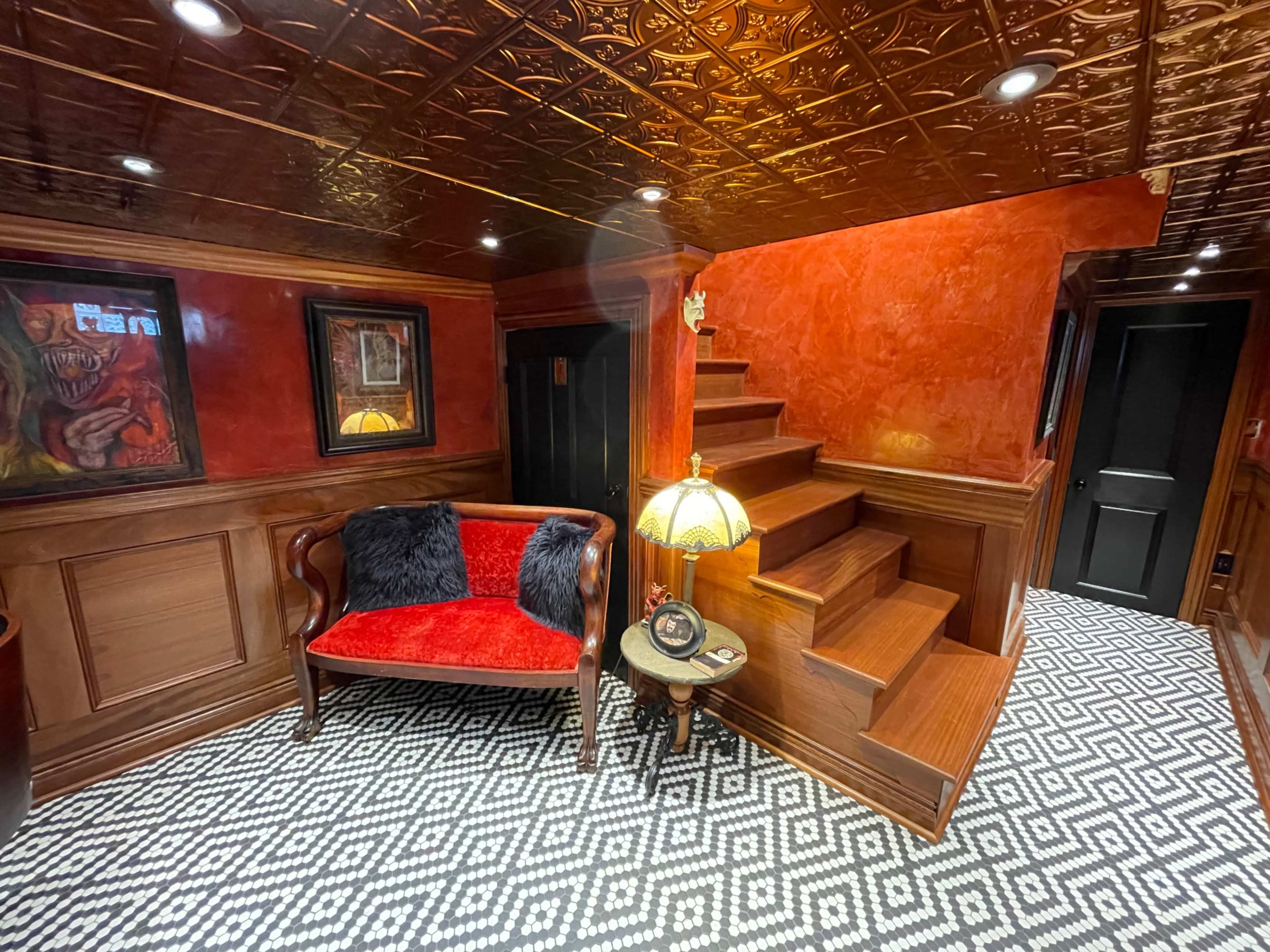 The image shows a richly decorated room with a red velvet couch, patterned black and white flooring, a staircase leading up, and a vintage lamp on a small table.