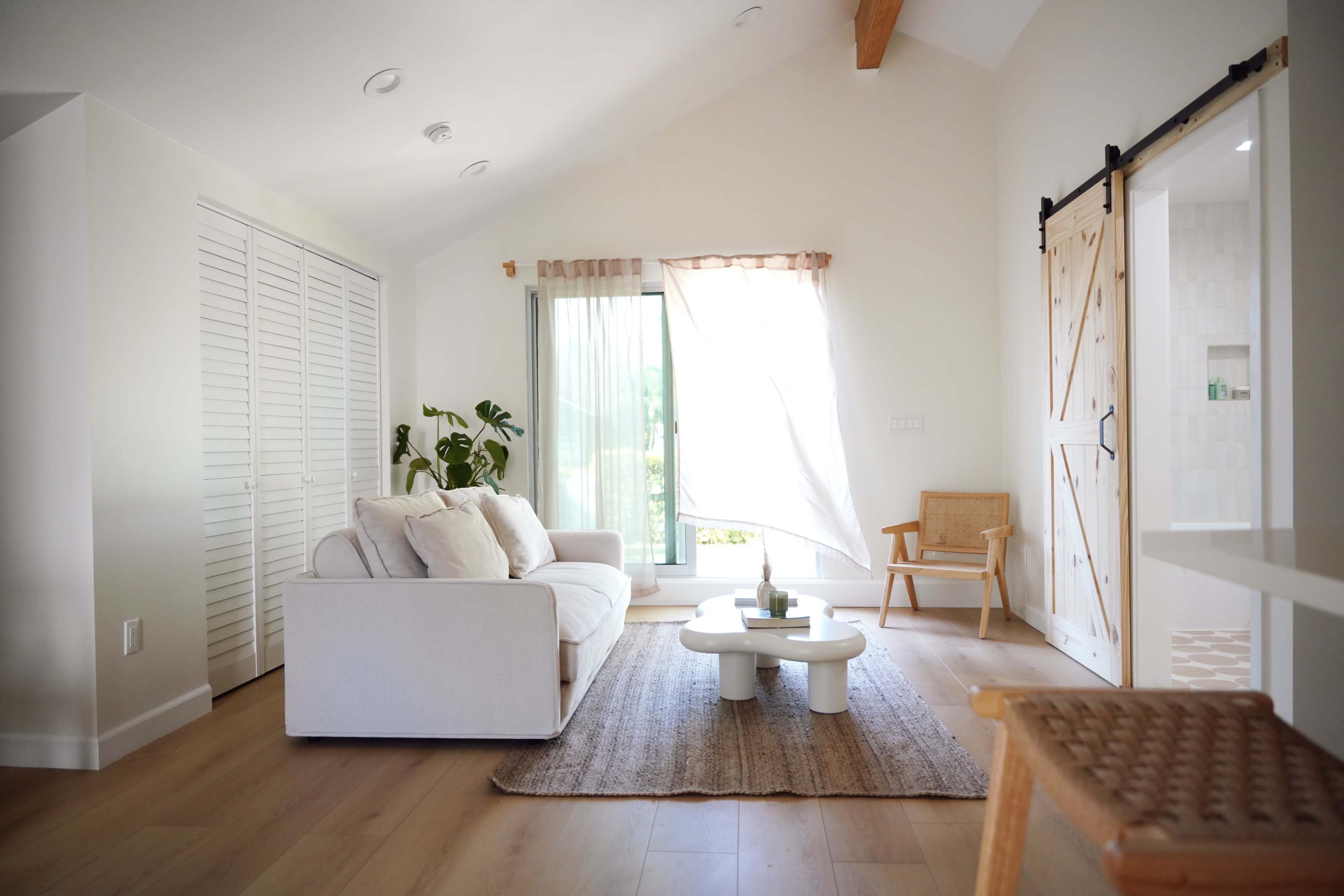 A bright, airy living room features a white sofa, a round coffee table, and large windows with sheer curtains, complemented by a woven rug and a wooden sliding door.