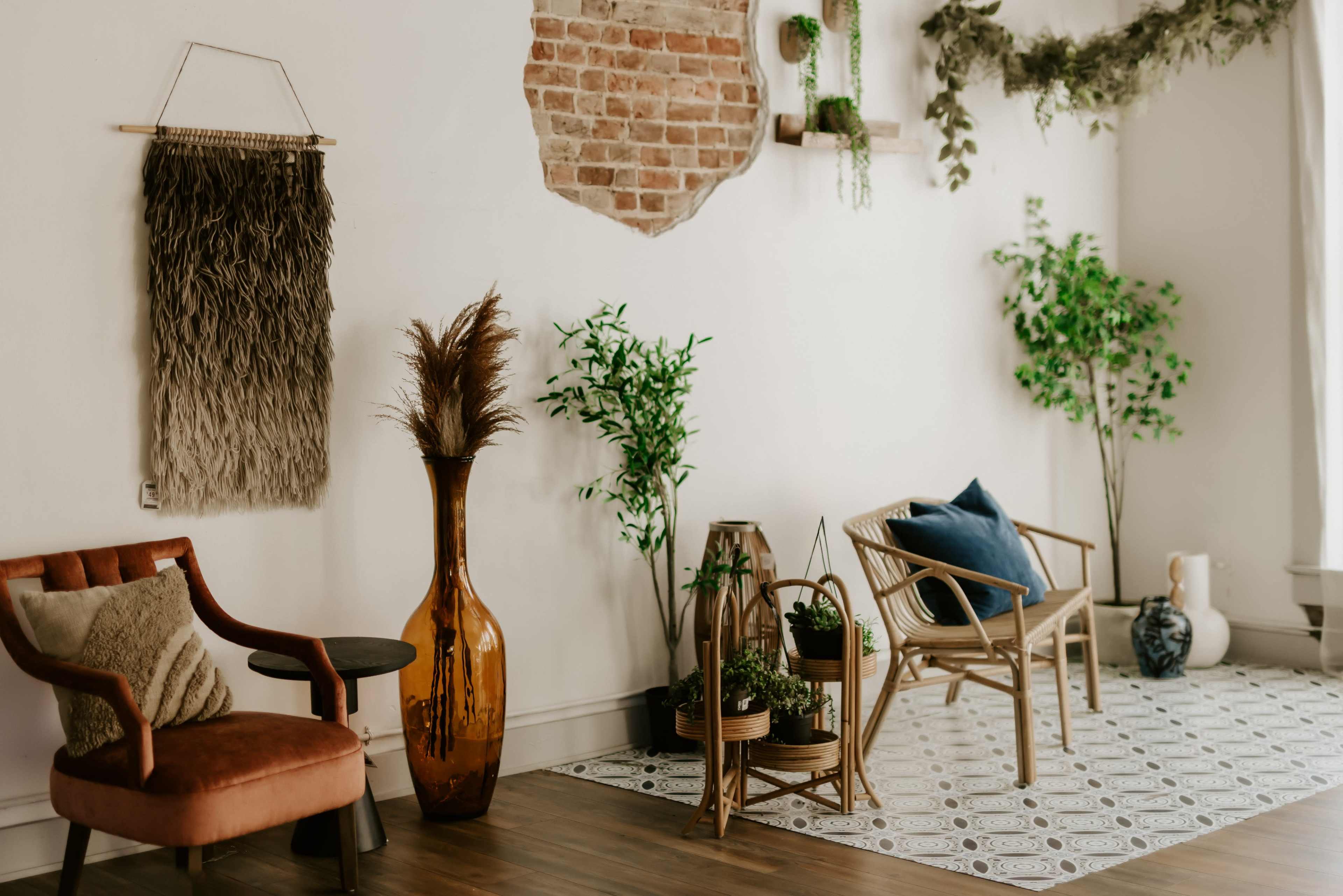 The image features a cozy interior space with a mixture of indoor plants, a stone wall, and stylish furniture including a rattan chair and an orange velvet armchair.