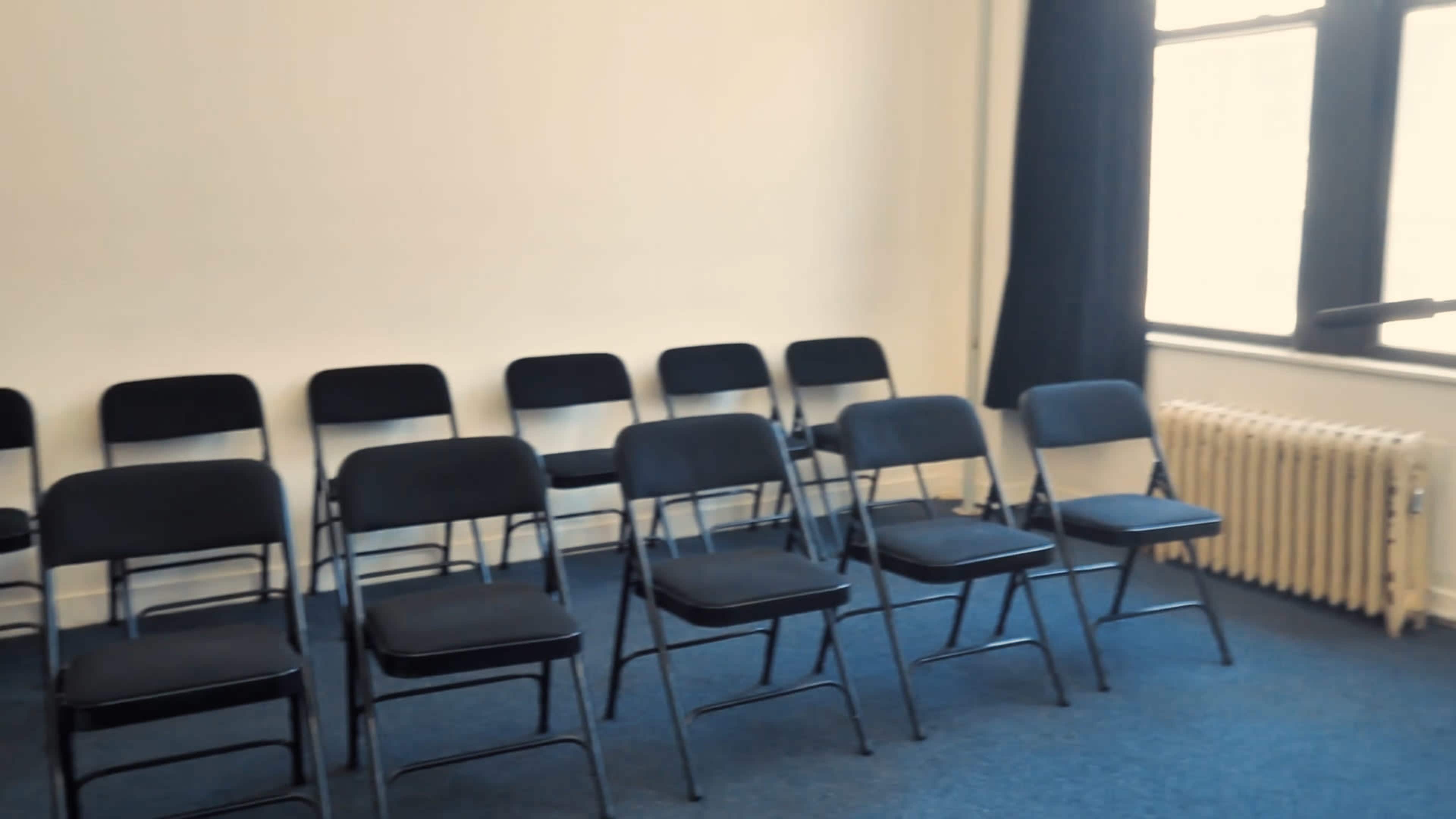 A row of black folding chairs is arranged in a room with a blue carpet and a window covered by a dark curtain.