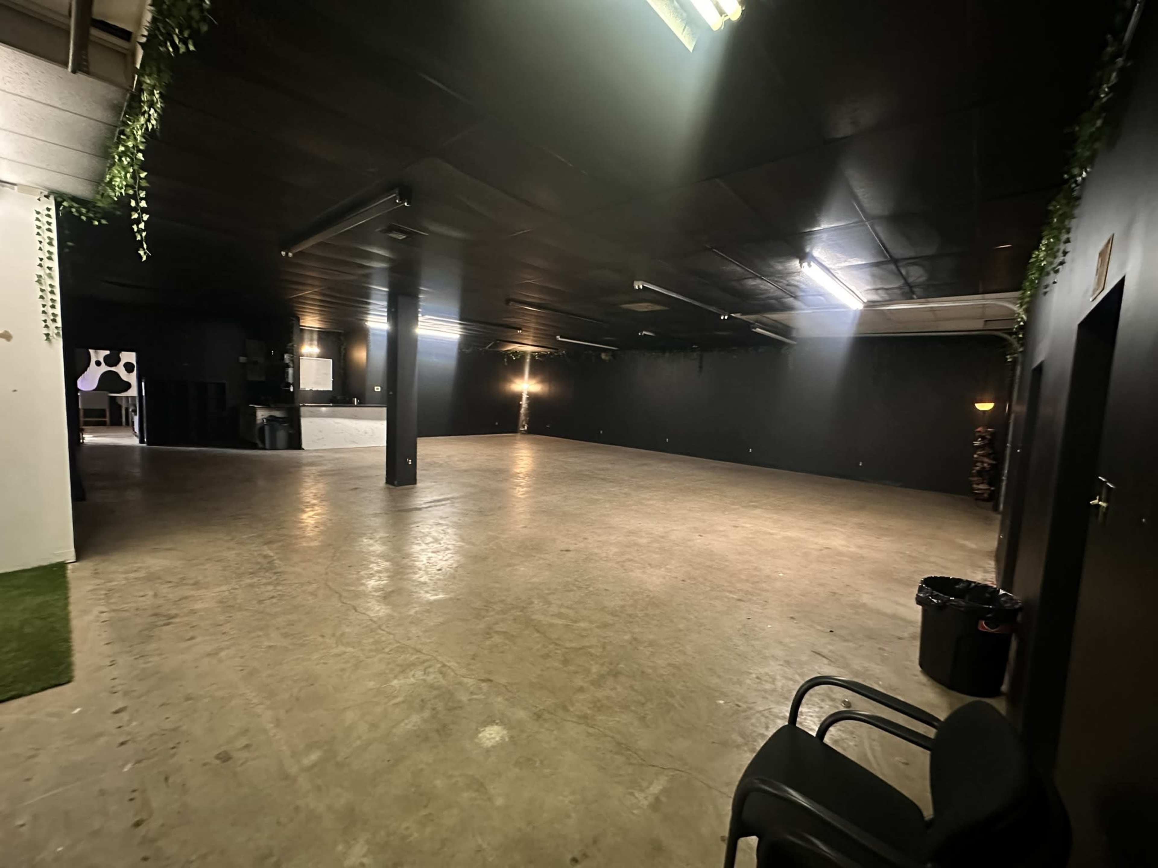 The image shows an empty industrial space with concrete flooring, black walls, and minimal furnishings, including a single chair and a trash can.