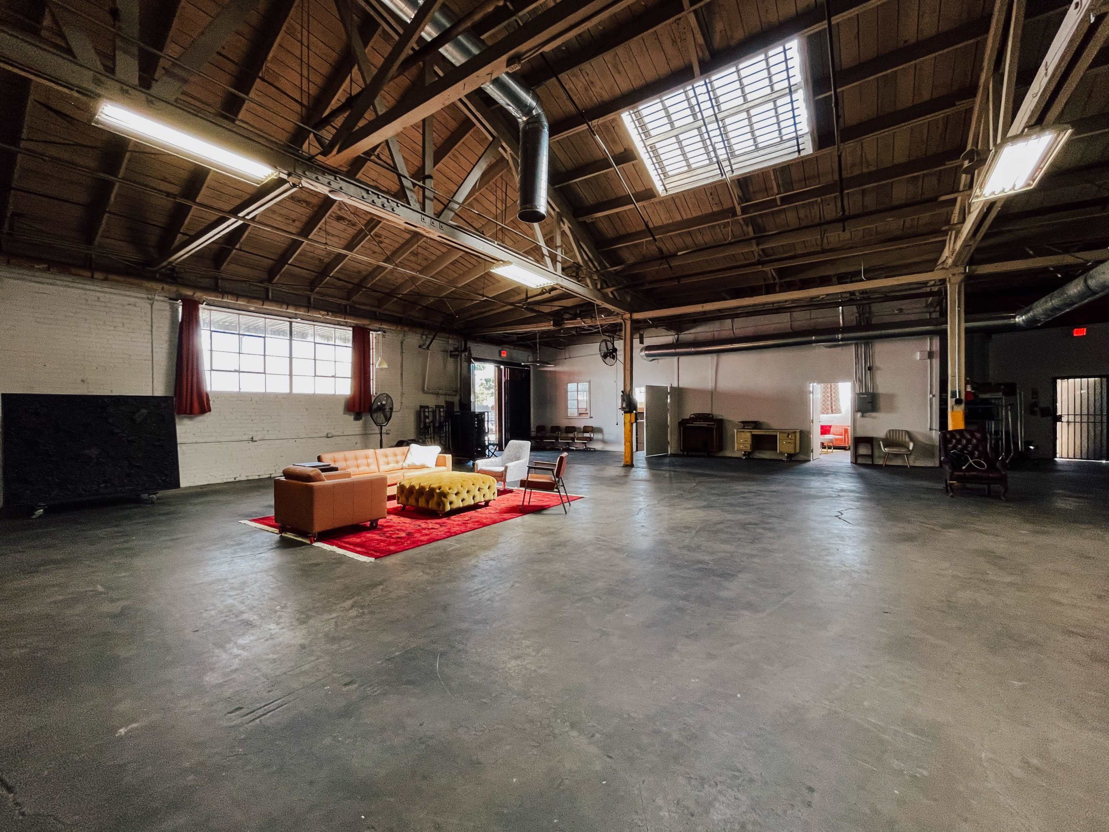 The image shows a spacious, industrial-style room with high wooden ceilings, large windows, and a seating area centered on a red rug.