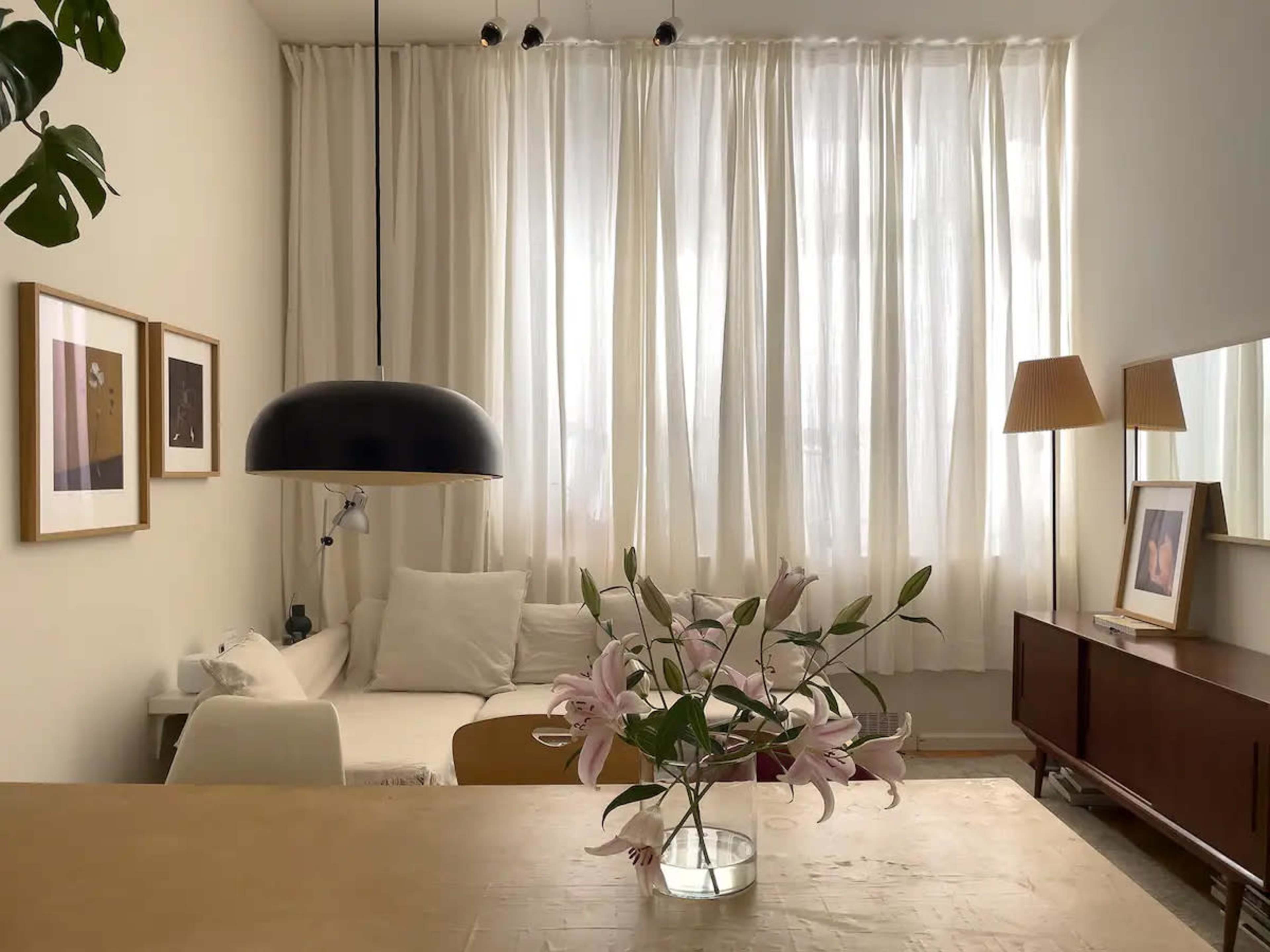 A cozy living room features a light-colored sofa, a wooden table with a vase of lilies, framed artwork on the walls, and large sheer curtains allowing natural light to enter.