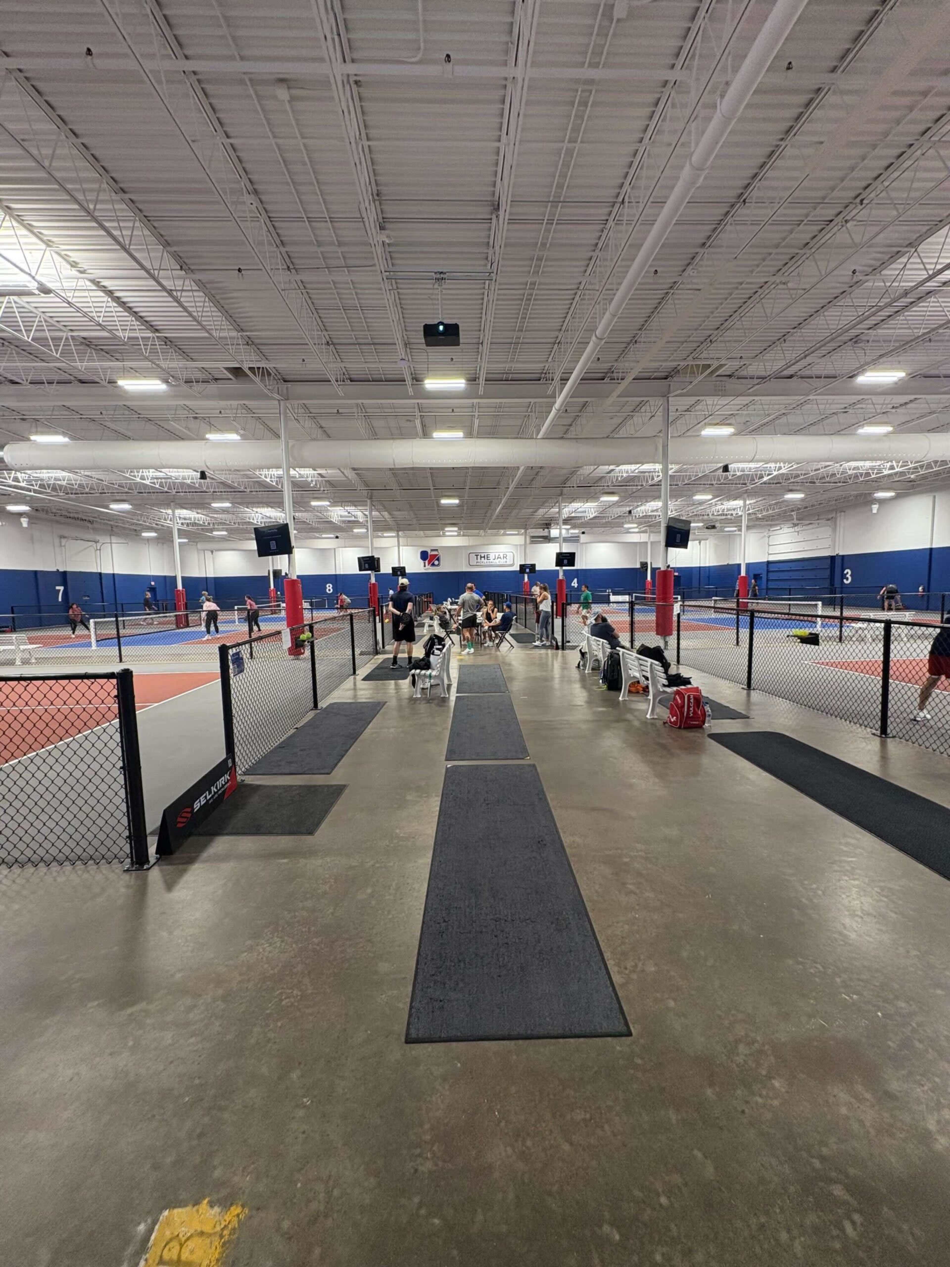 The image shows an indoor tennis facility with multiple courts, players engaged in matches, and seating areas on either side.