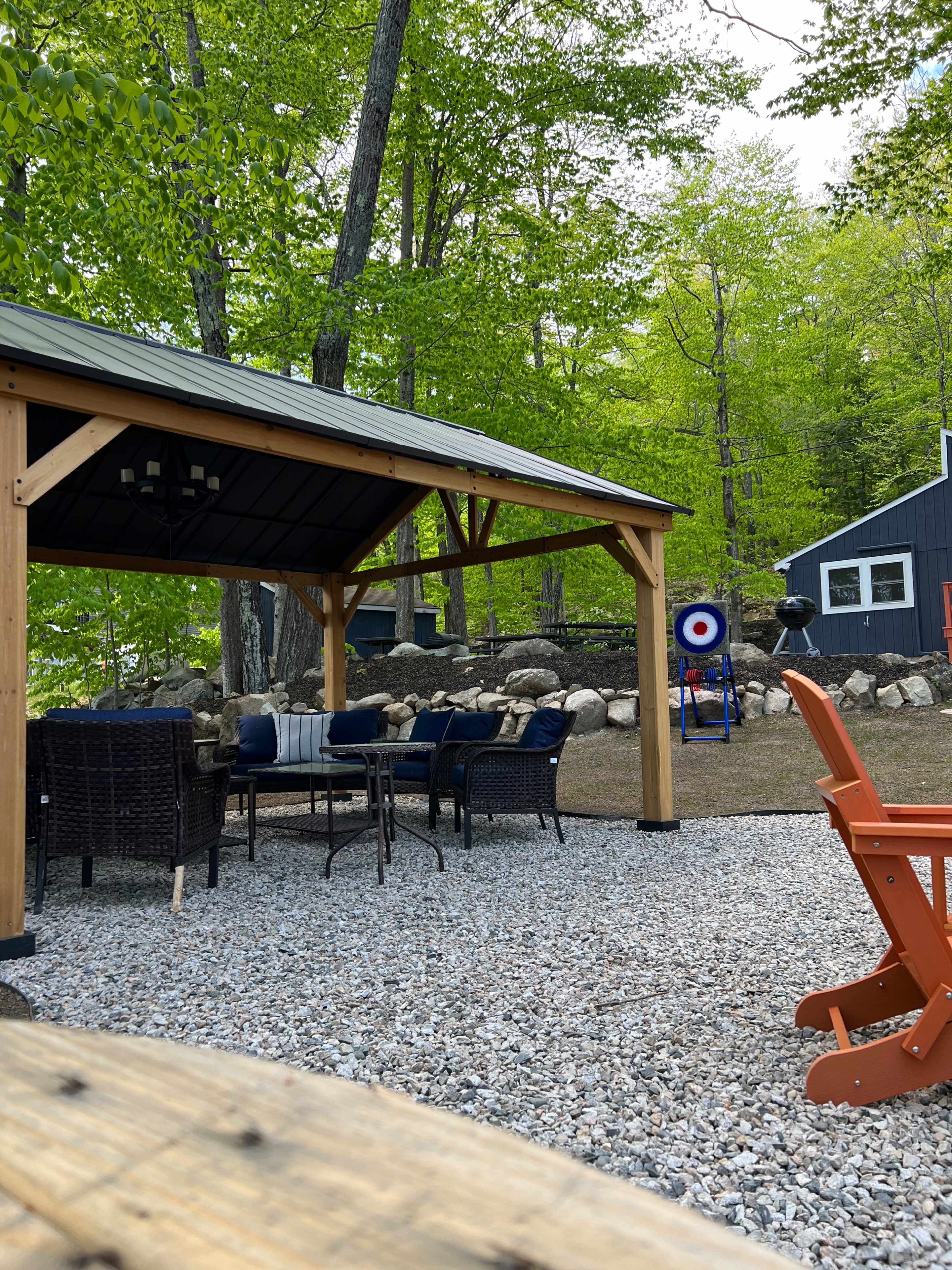 A gravel patio area features a covered shelter with seating, surrounded by trees and a target set up in the background.