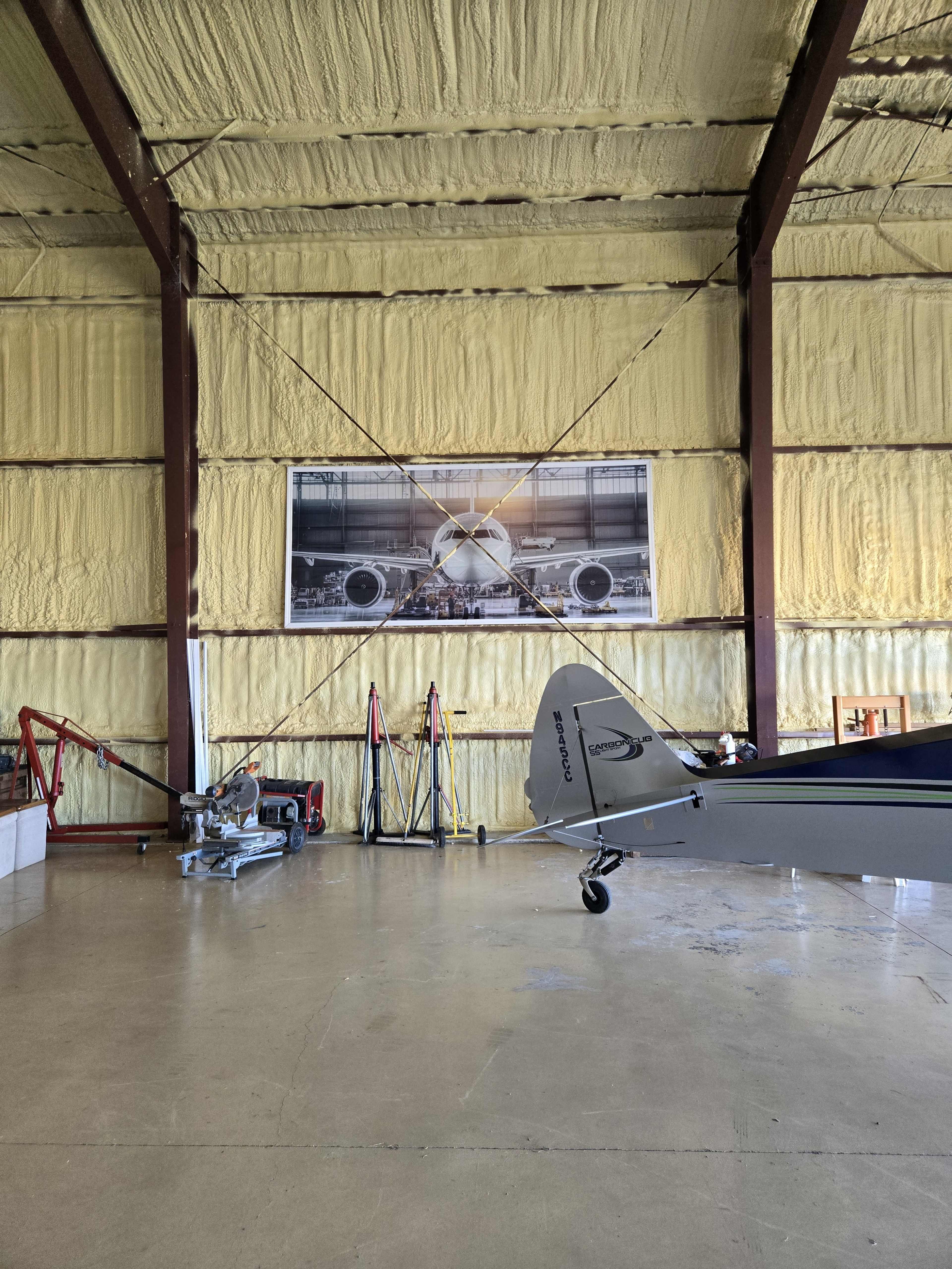The image shows a hangar with a small aircraft in the foreground, large aircraft prints on the wall, and various tools and equipment along the side.