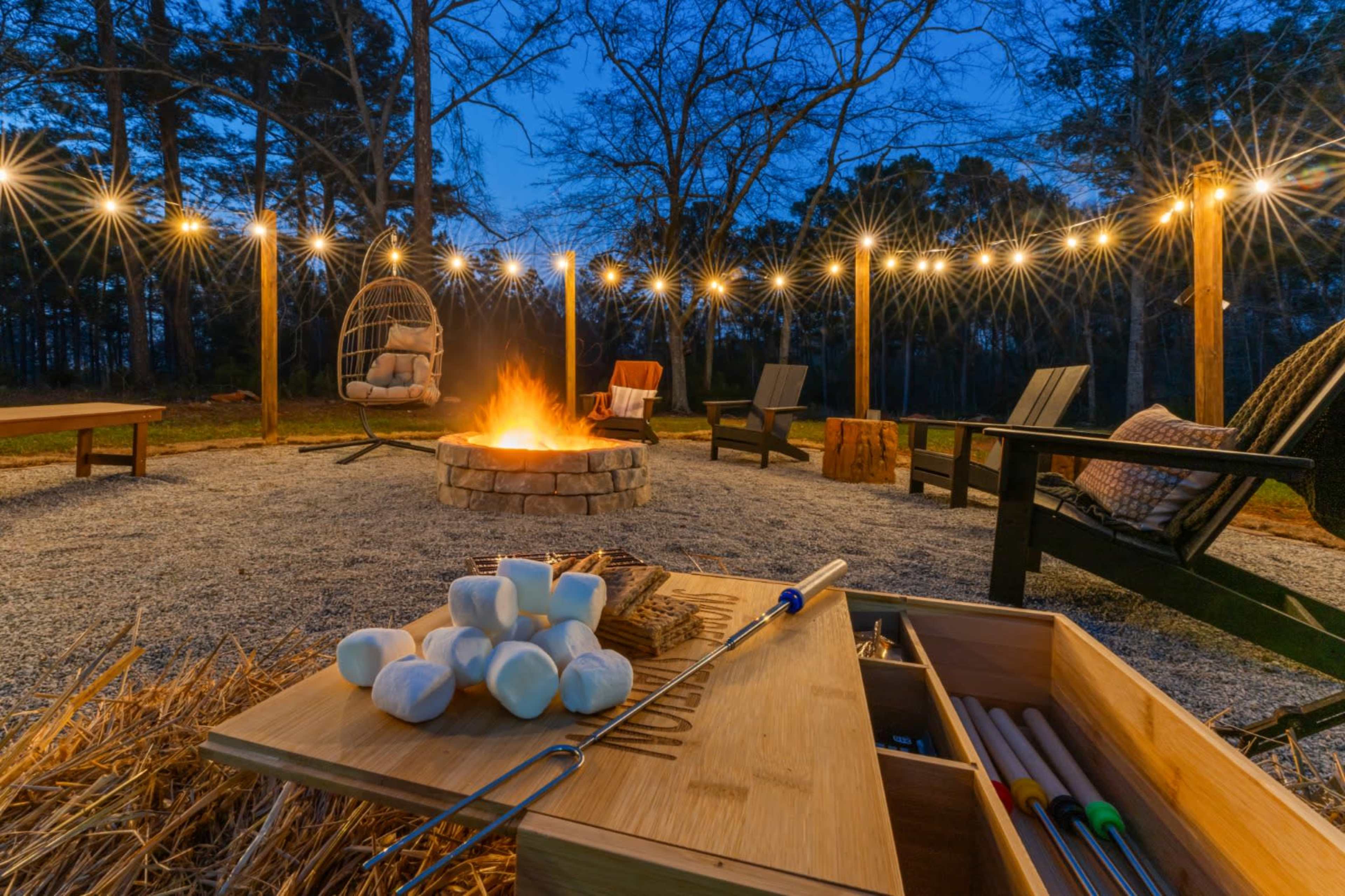A cozy outdoor campfire scene at dusk features a circular stone fire pit surrounded by wooden chairs and string lights illuminating the area.
