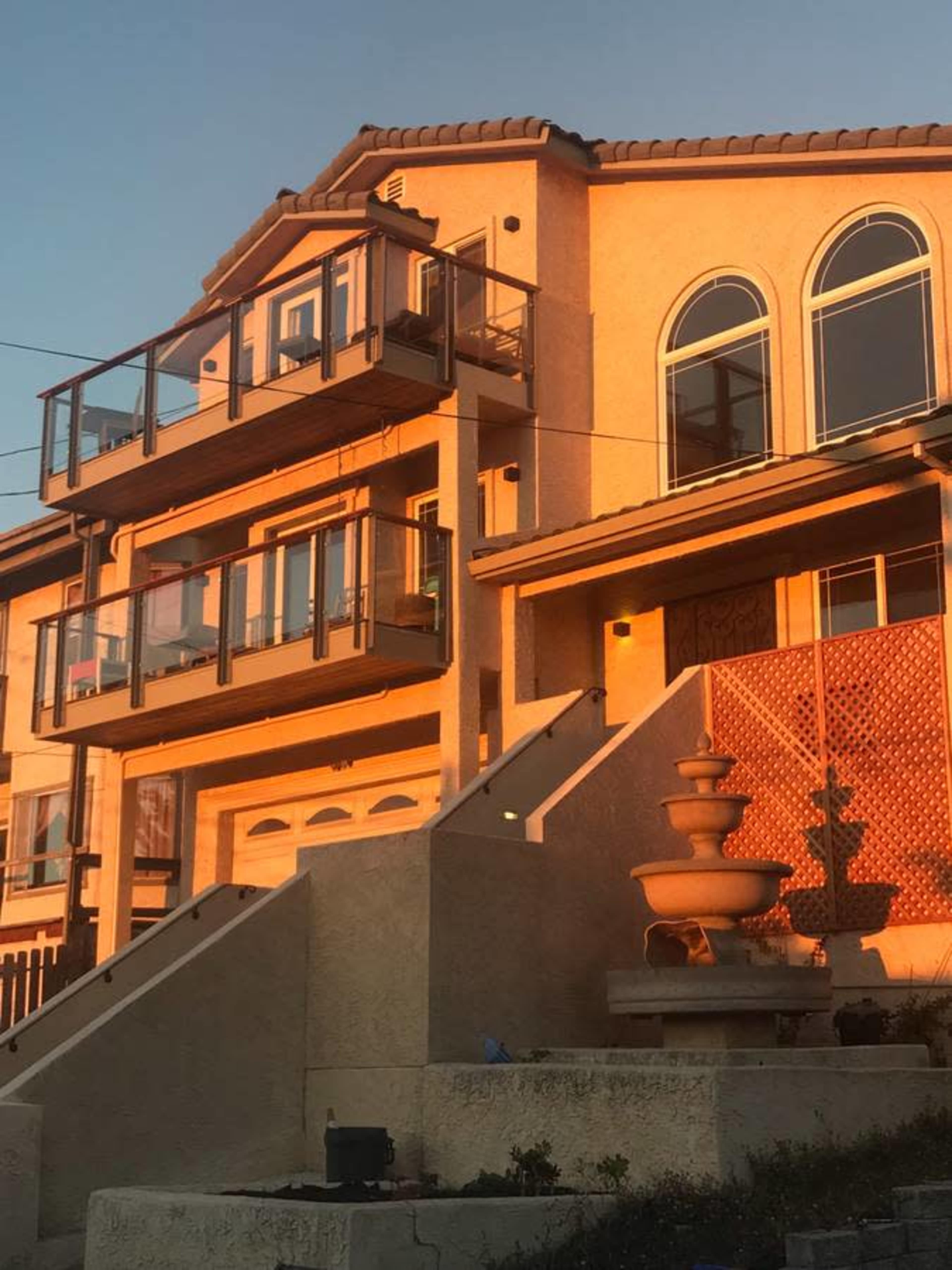 A multi-story house with balconies and a fountain at the foot of its steps is illuminated by the setting sun.