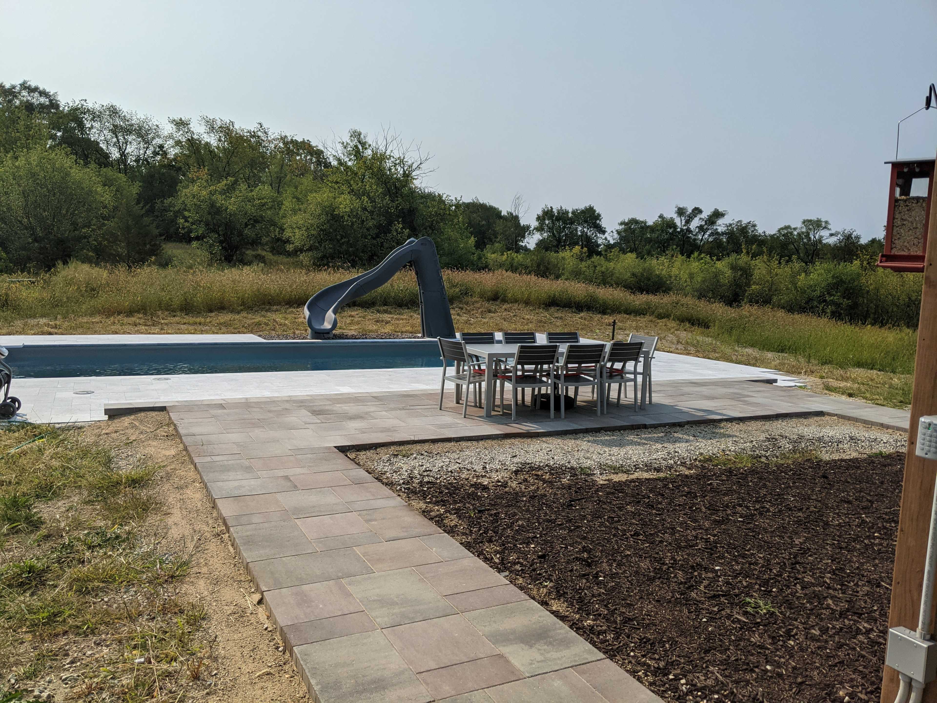 A swimming pool with a water slide next to a patio featuring a dining table and chairs, surrounded by grassy areas and trees.