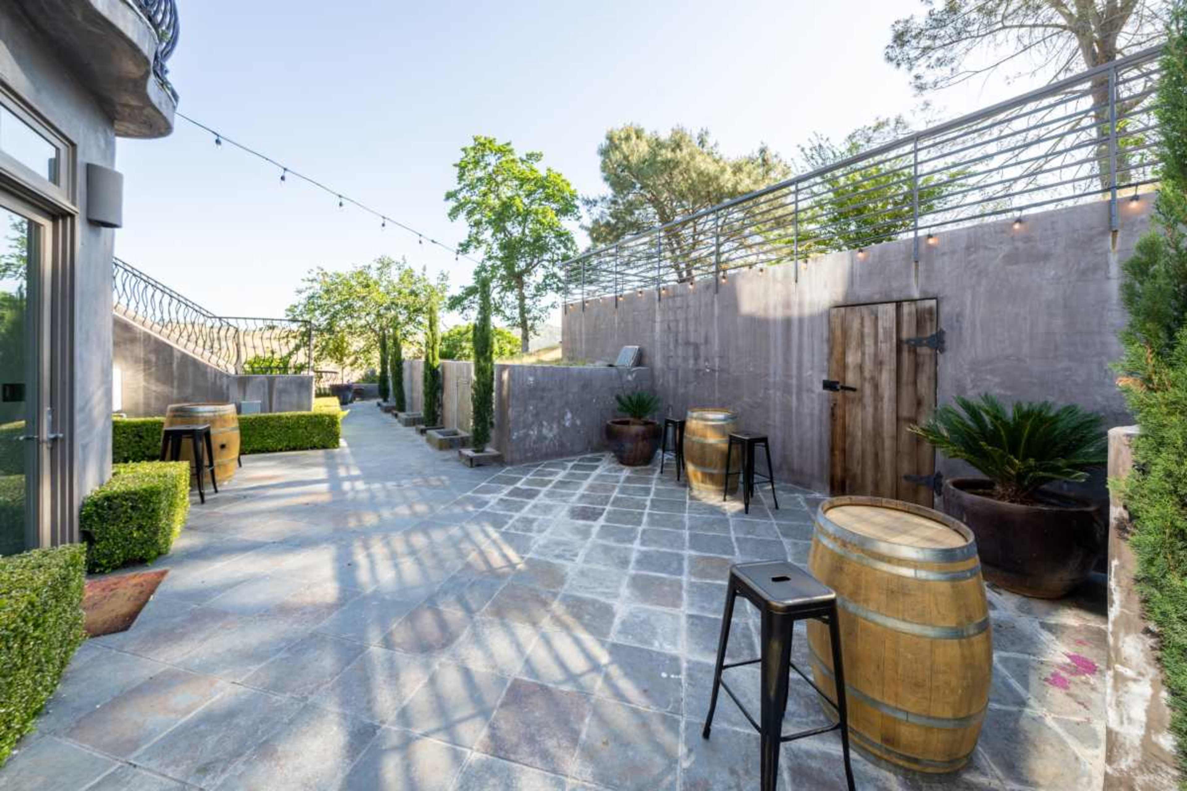 The image shows a spacious outdoor patio area with stone flooring, wooden barrels, and greenery, bordered by a low wall and a wooden door.