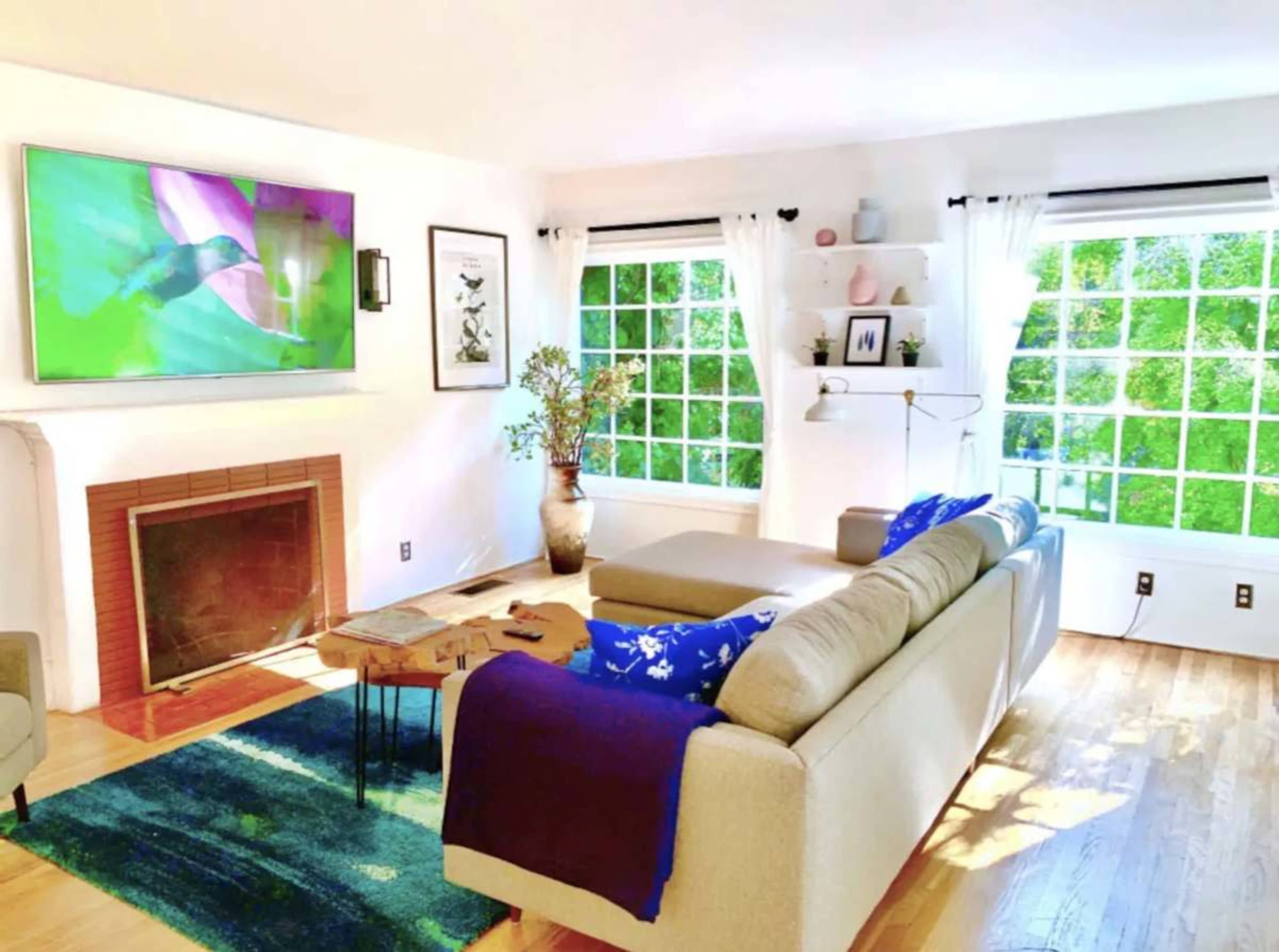 The living room features a beige sectional sofa, a coffee table, a fireplace, and large windows with sheer white curtains letting in natural light.