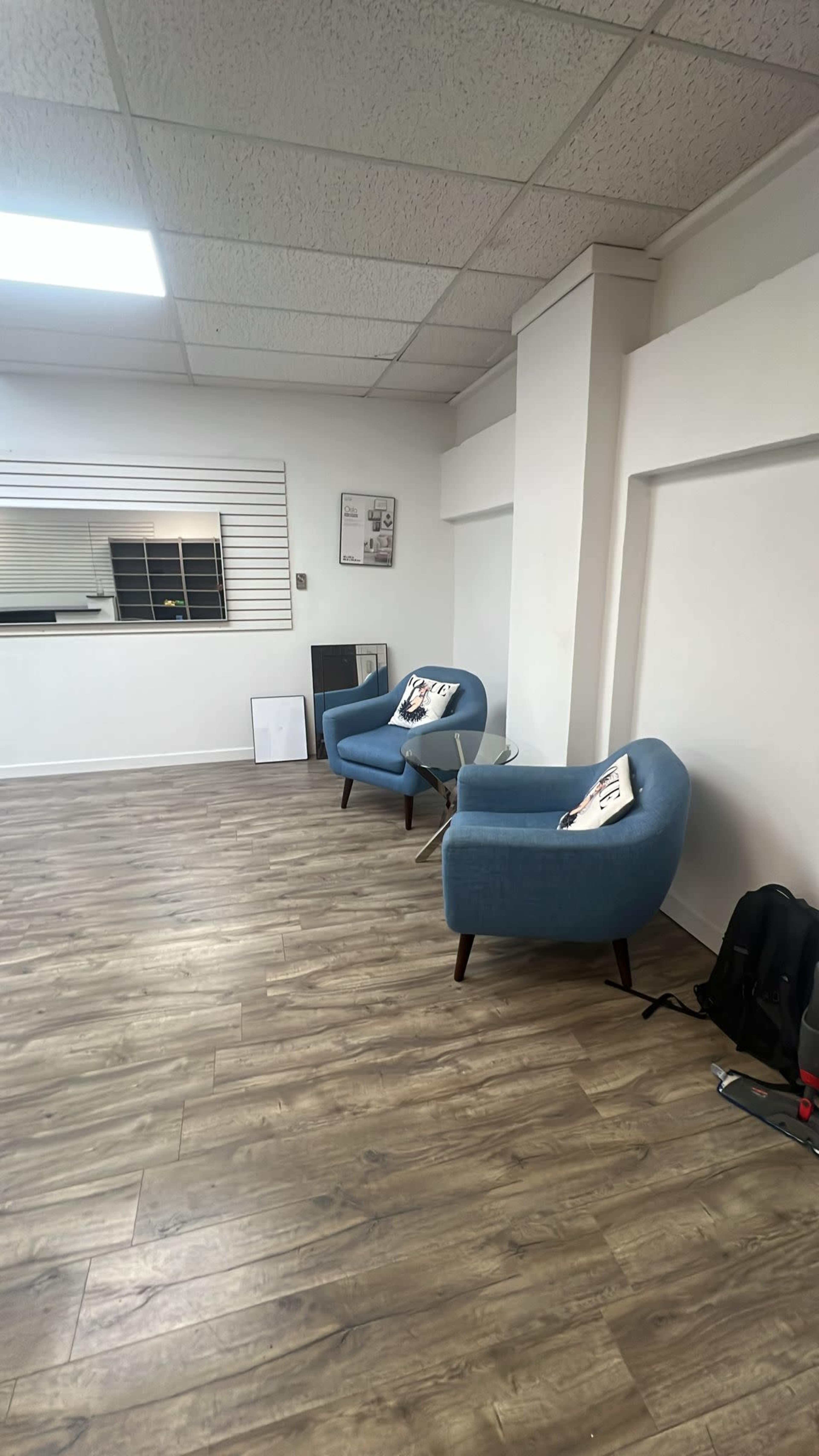 The image shows an empty room with light wooden floors, two blue chairs with decorative pillows, and a window with horizontal blinds.