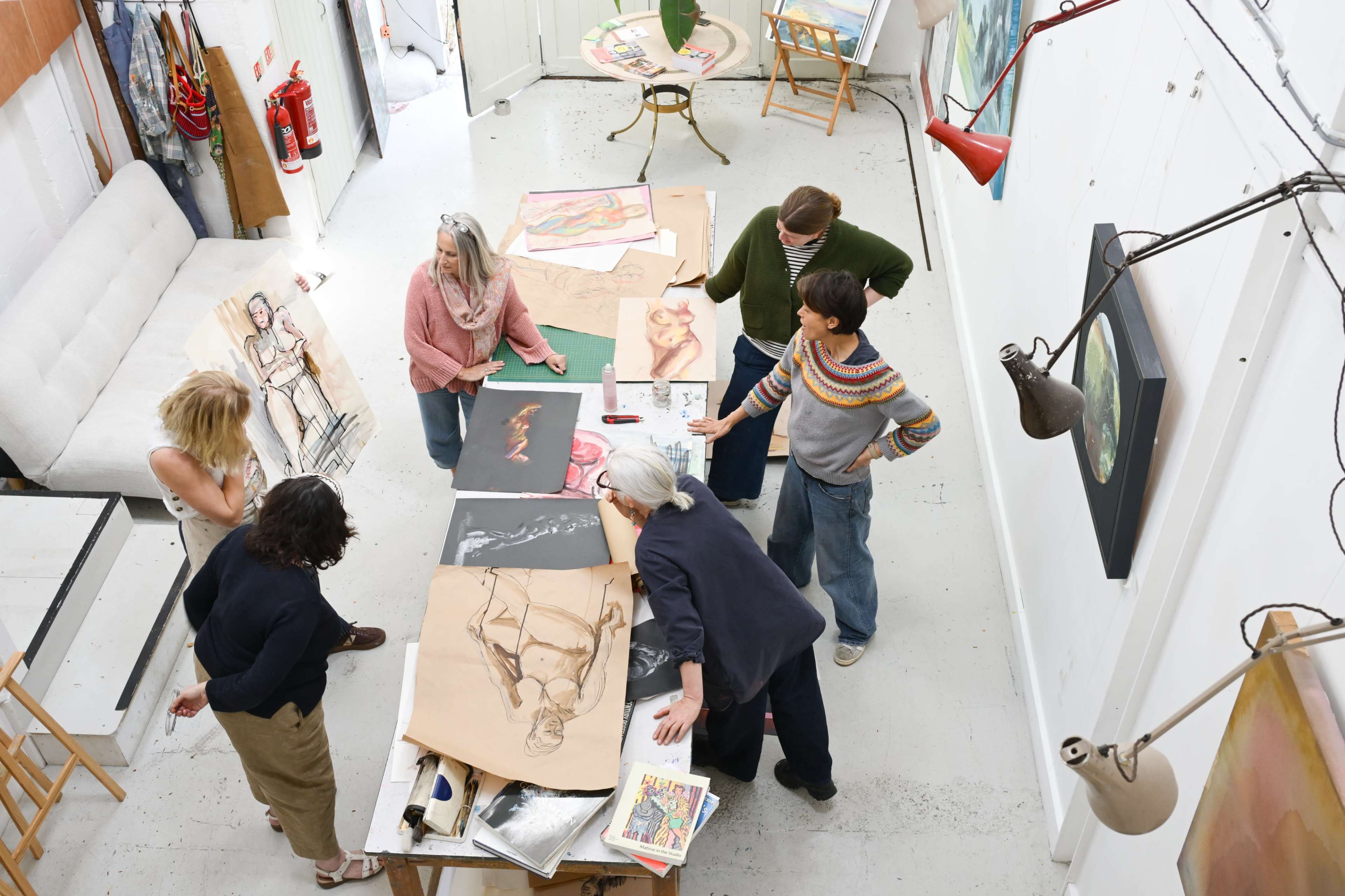 A group of seven people is gathered around a table in an art studio, examining various drawings and artworks spread out across the surface.