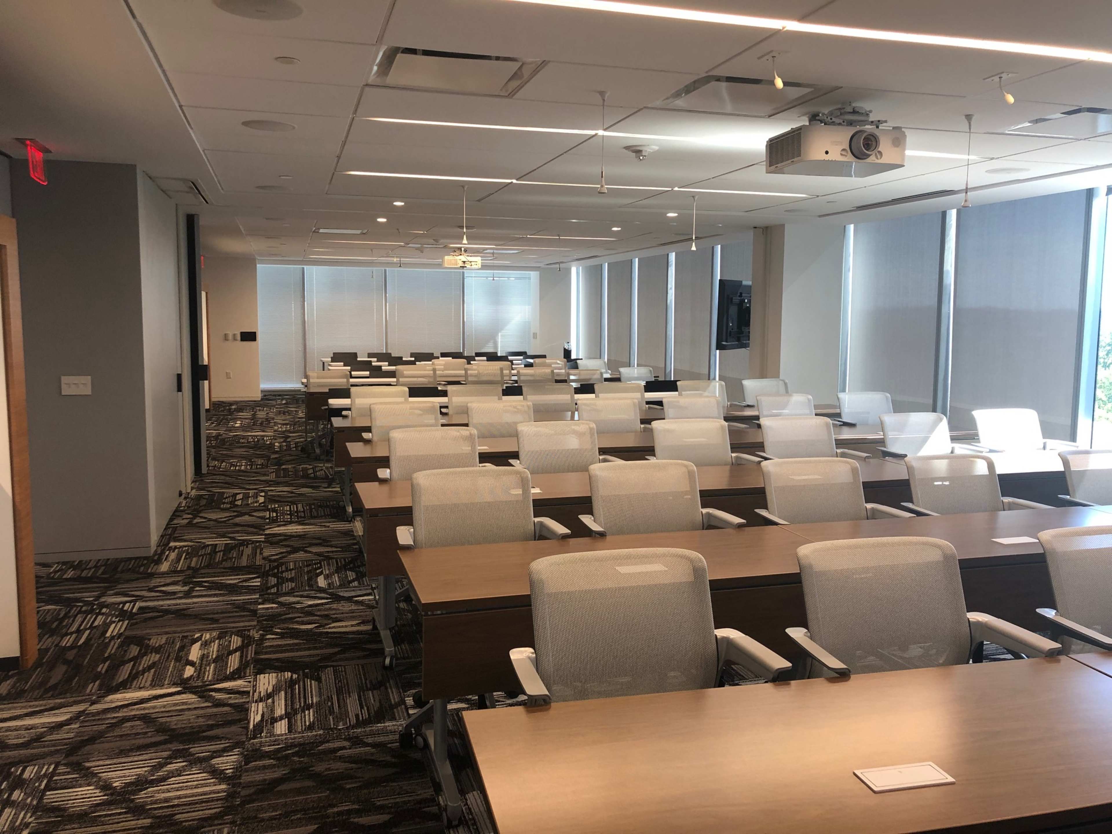 A modern conference room is set up with rows of tables and chairs, with large windows allowing natural light and a projector mounted on the ceiling.