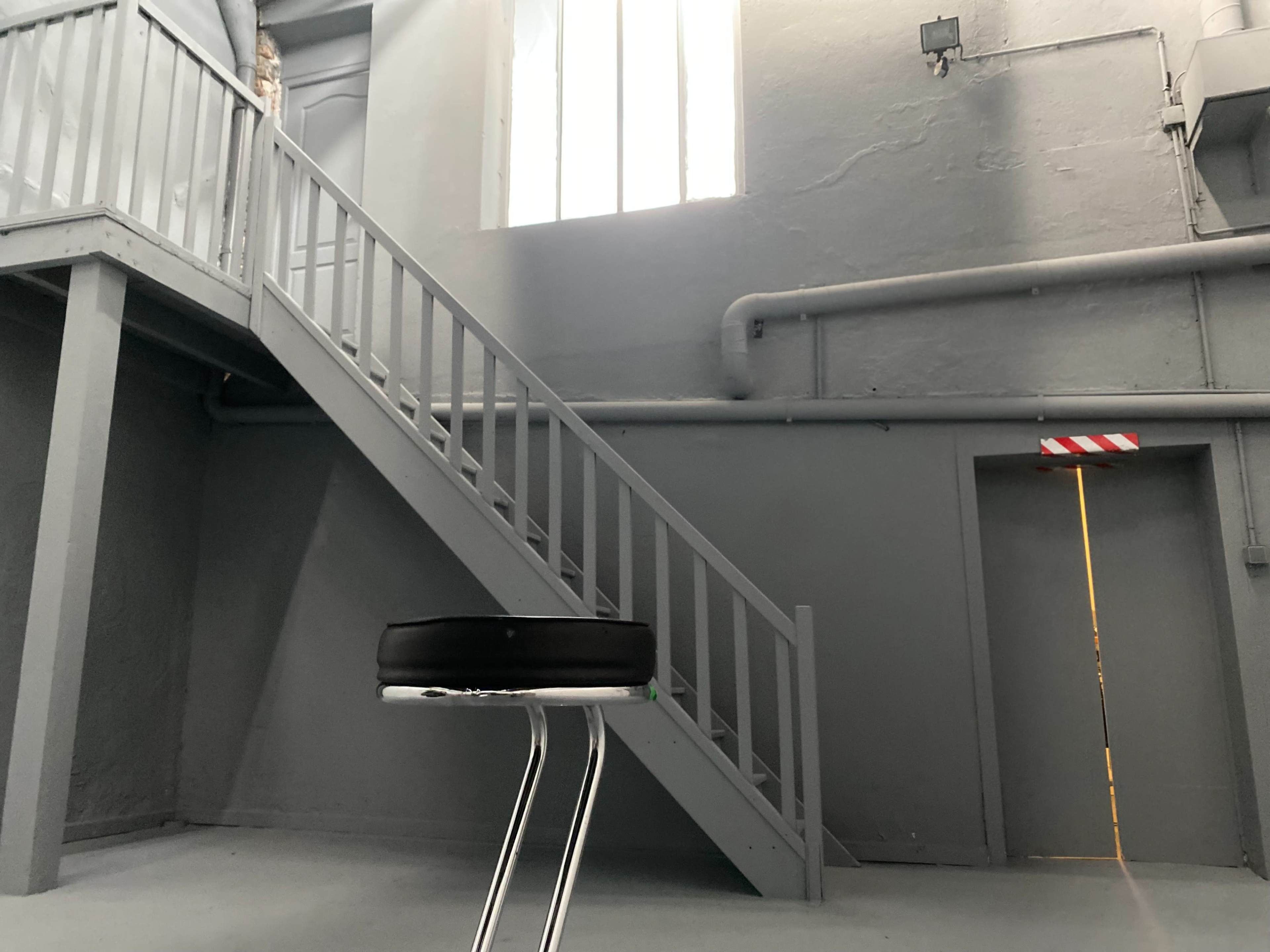 A black stool is positioned in an empty, gray room with a staircase leading to a door illuminated by light at the top.