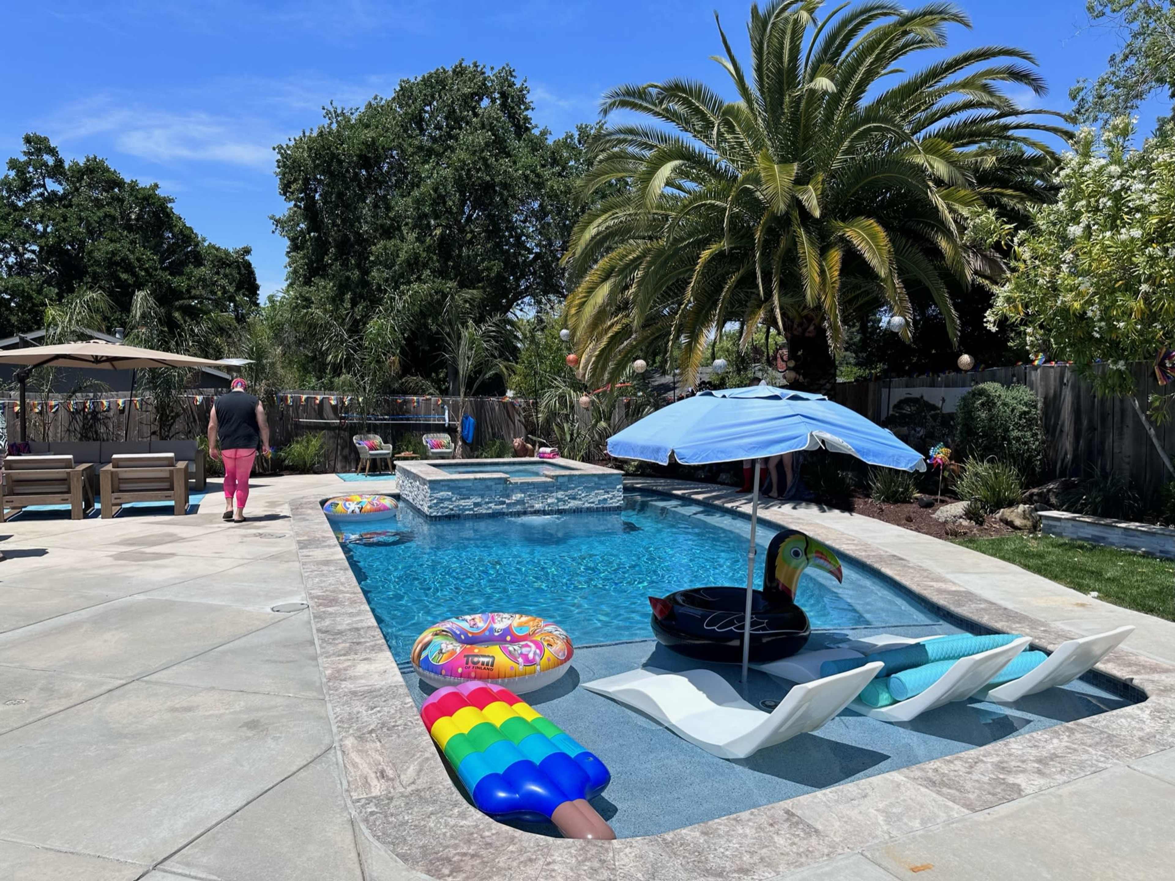 The image shows a residential backyard with a swimming pool surrounded by lounge chairs, inflatable pool toys, and palm trees under a clear blue sky.