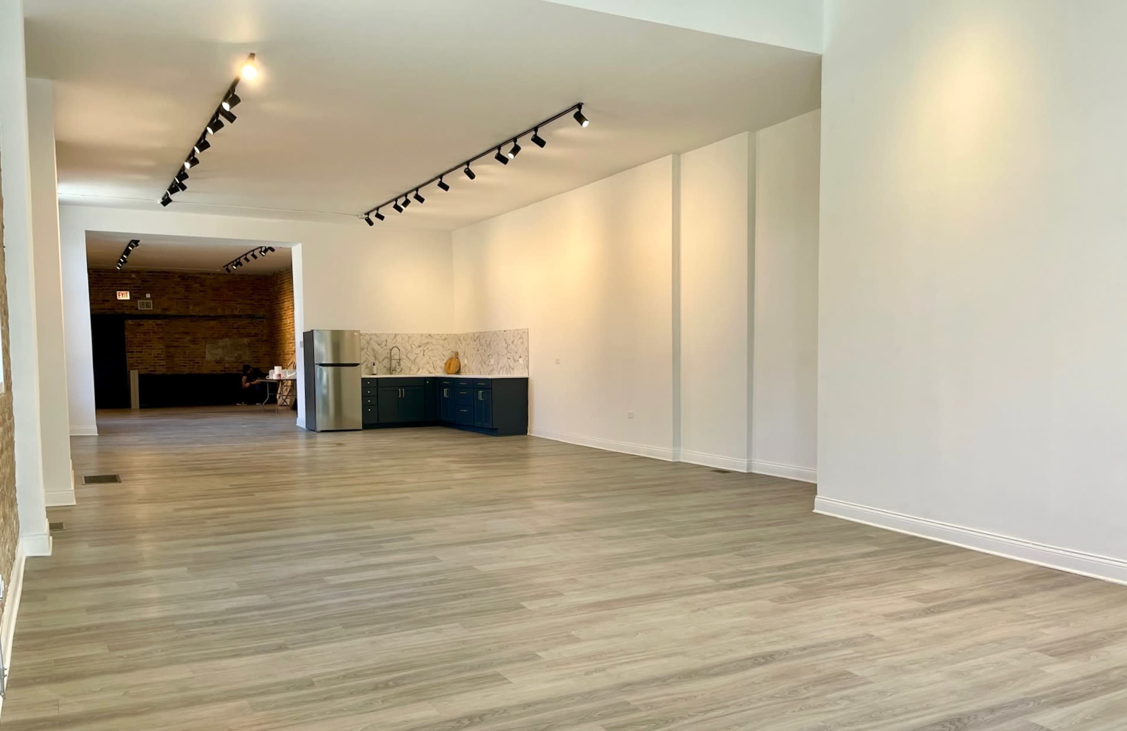 A spacious, empty room with light-colored walls and a wooden floor, featuring a kitchen area with blue cabinets at one end.