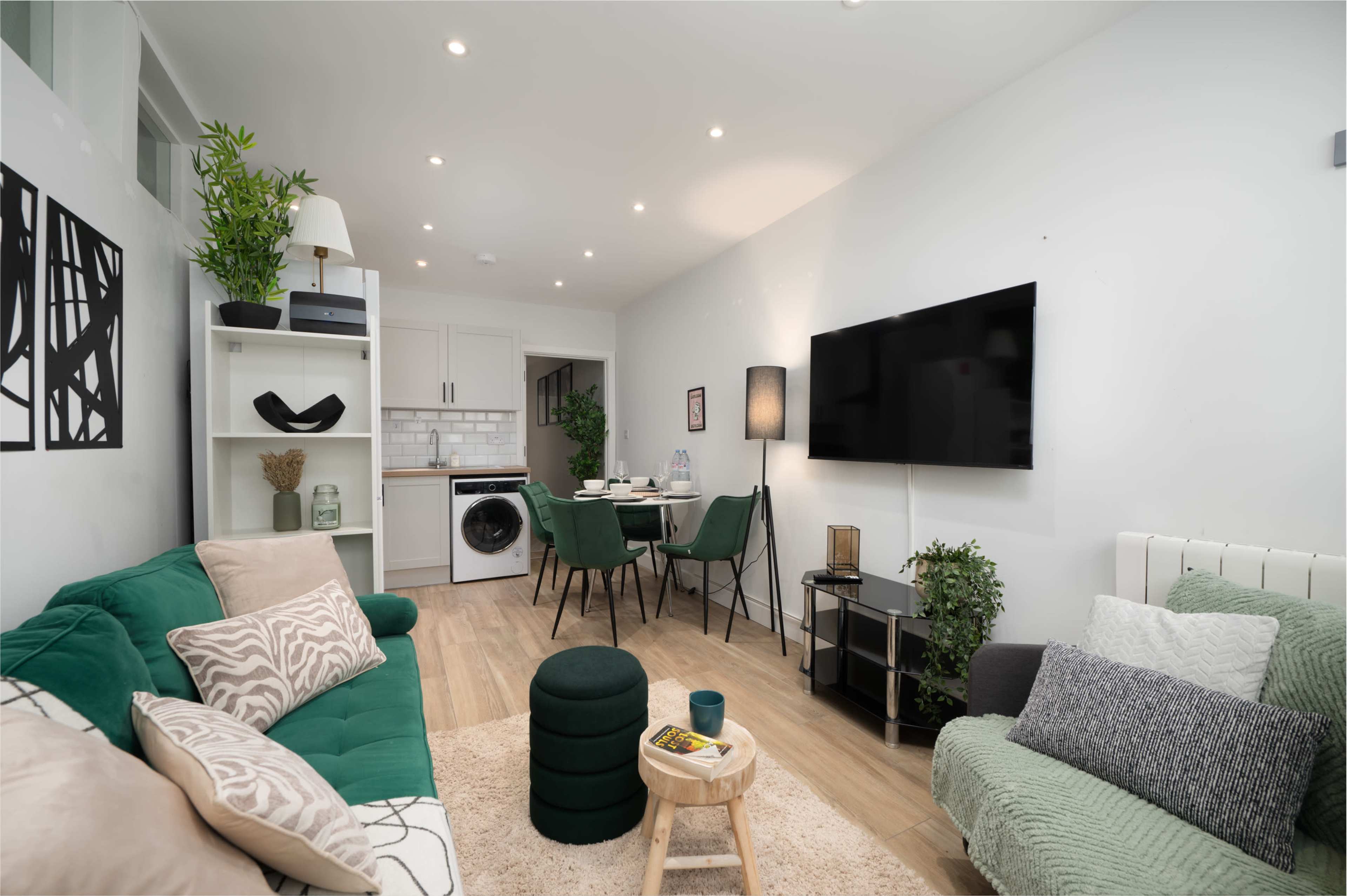 The image shows a modern living room featuring green sofas, a dining area with four chairs, and a wall-mounted television.