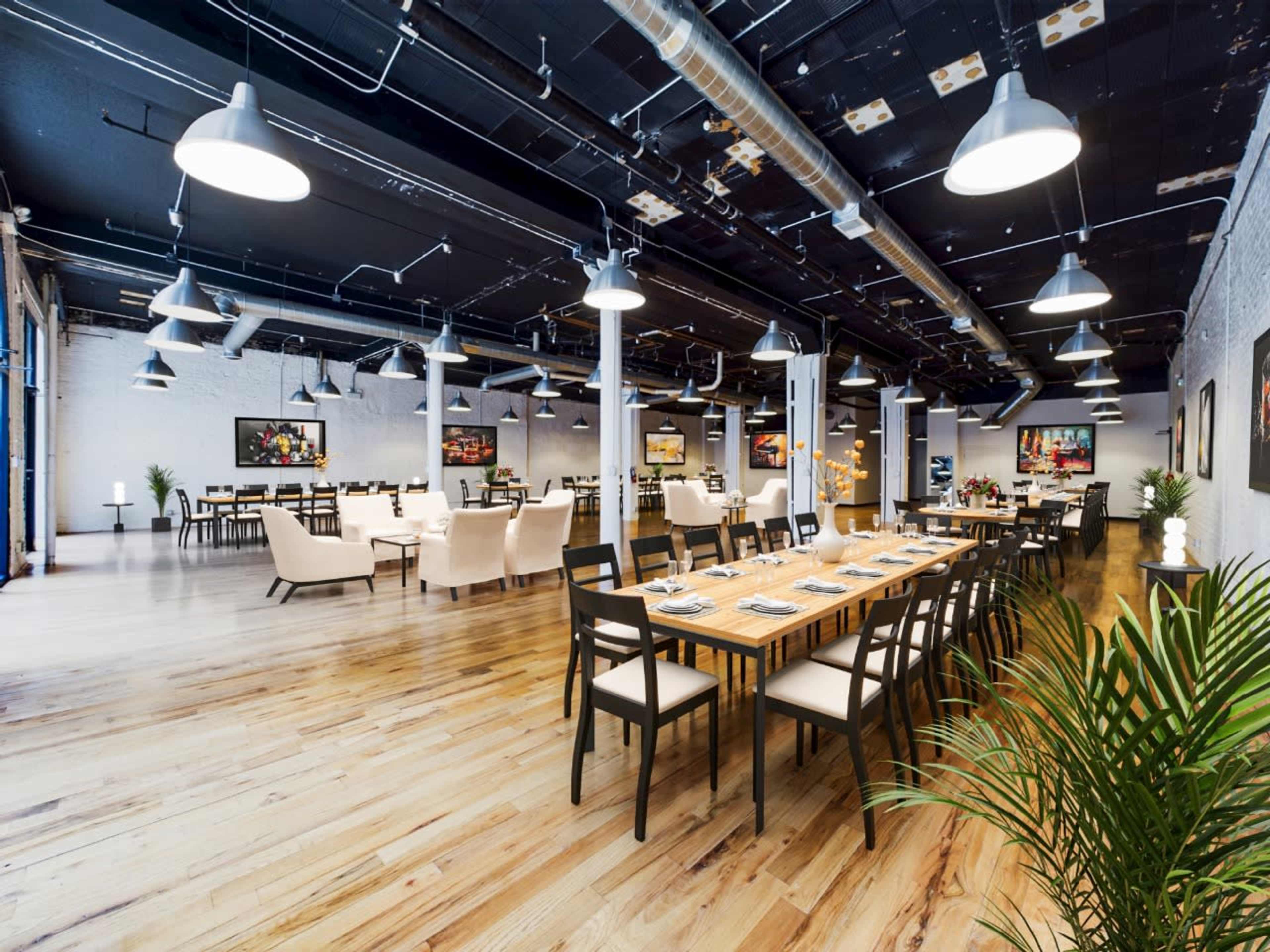 A spacious dining area with wooden flooring, long tables set for dining, and soft lighting fixtures suspended from the ceiling.