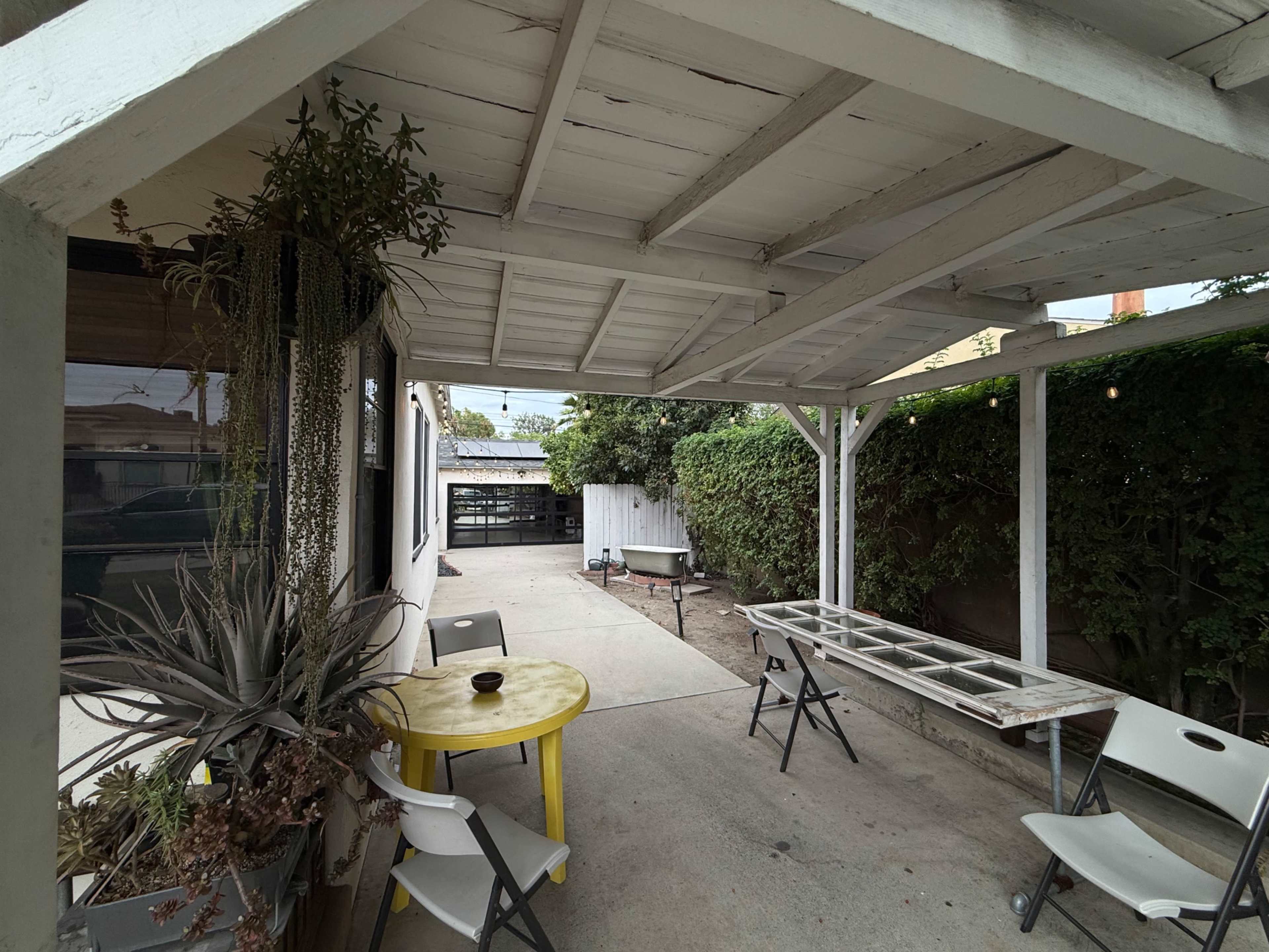 The image shows a covered outdoor patio area with a yellow table and several chairs, surrounded by greenery and a concrete floor.