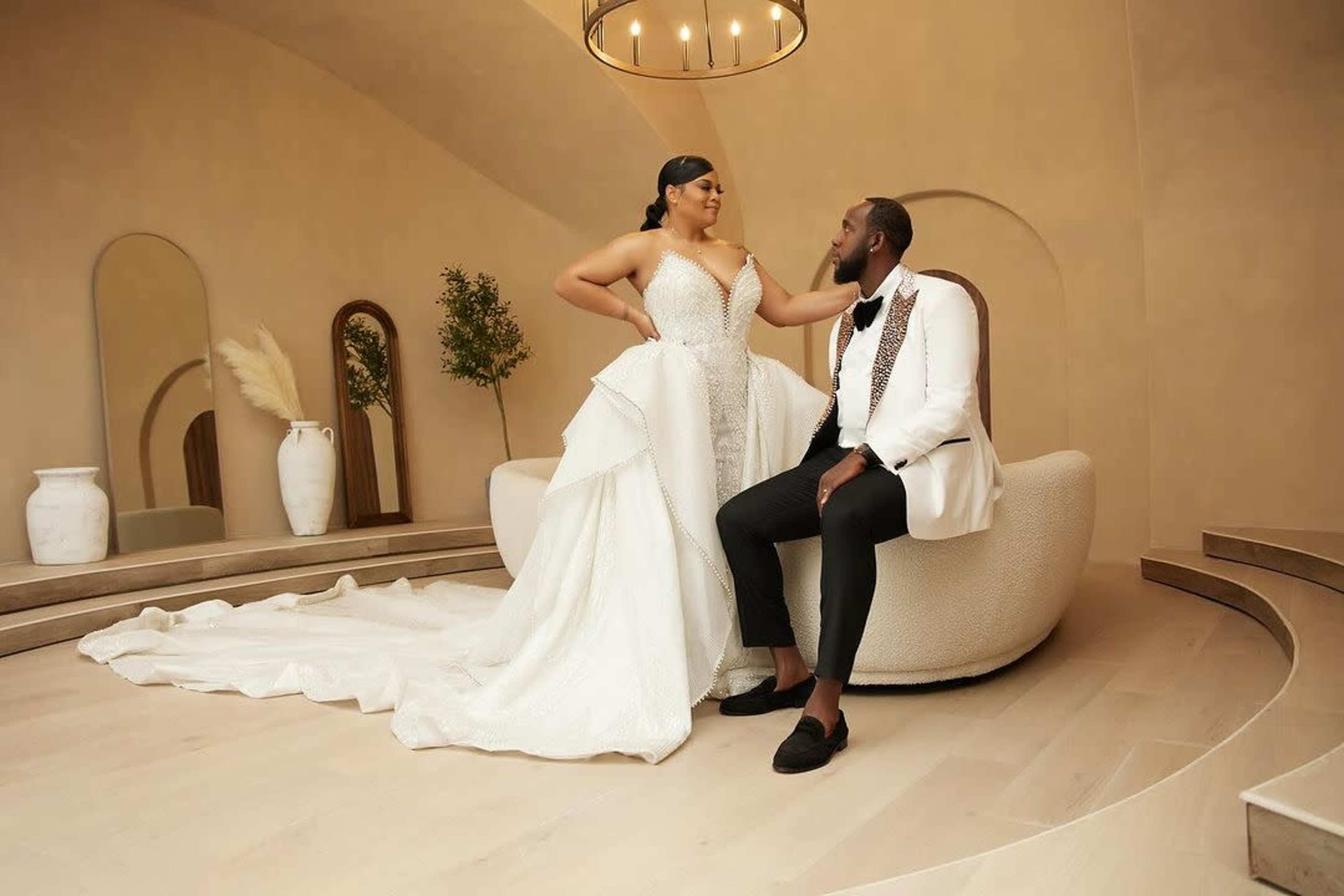 A bride in a flowing white gown sits next to a groom in a black tuxedo on a round couch in a softly lit modern room with curved walls and decorative mirrors.
