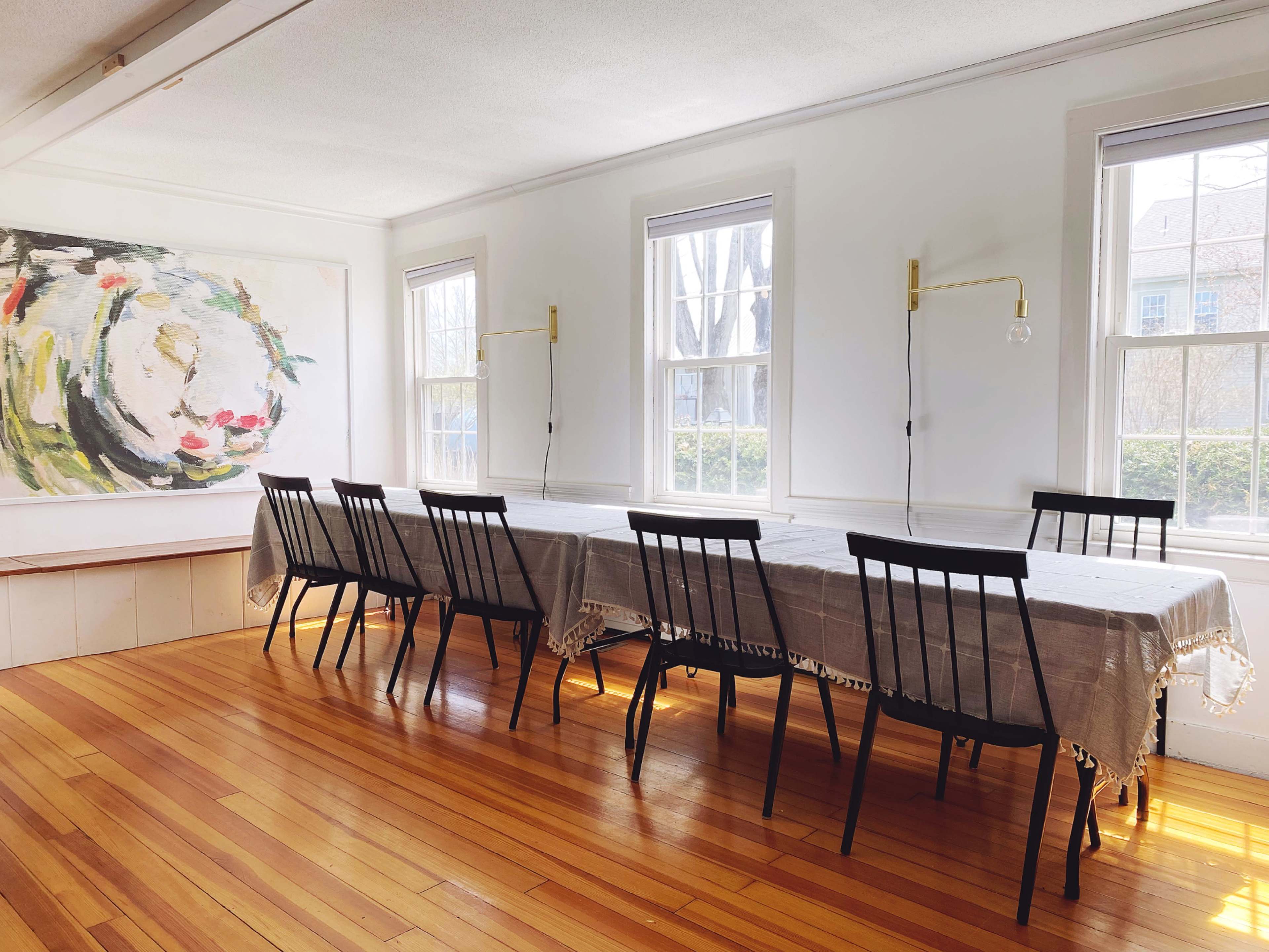 A long table with a gray tablecloth and eight black chairs is positioned in a well-lit dining room featuring large windows and a colorful painting on the wall.