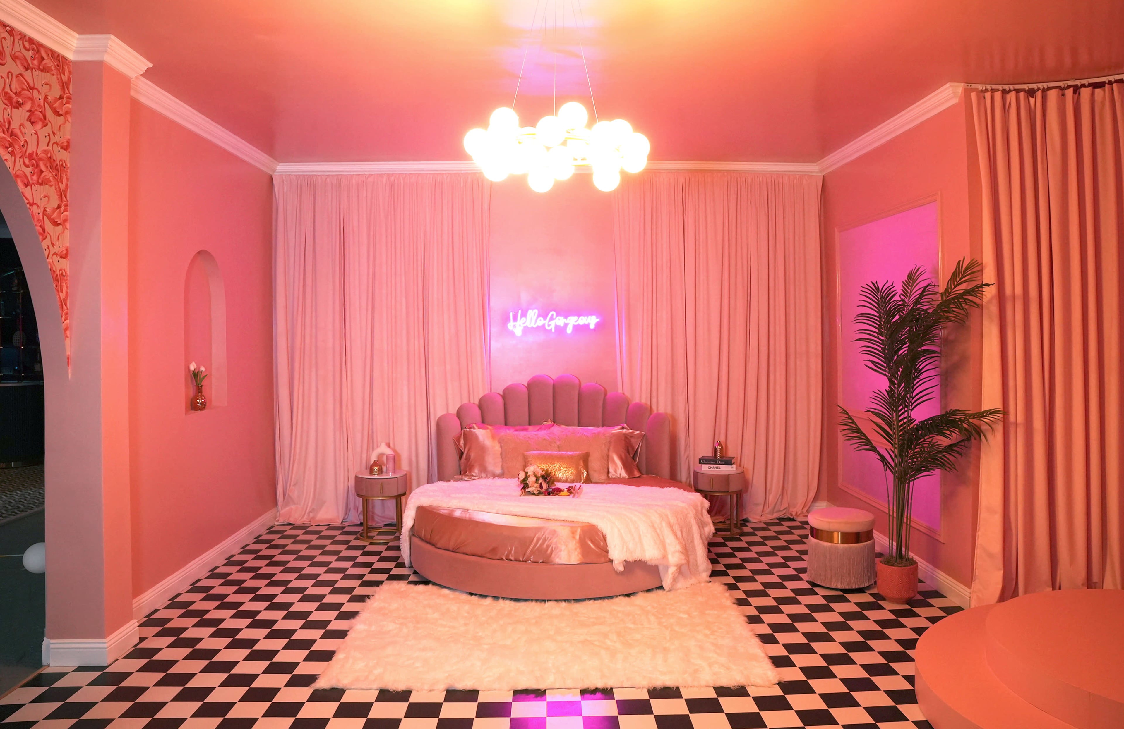 A pink-themed room with a round bed, stylish lighting, and a checkered floor, accentuated by curtains and a neon sign.