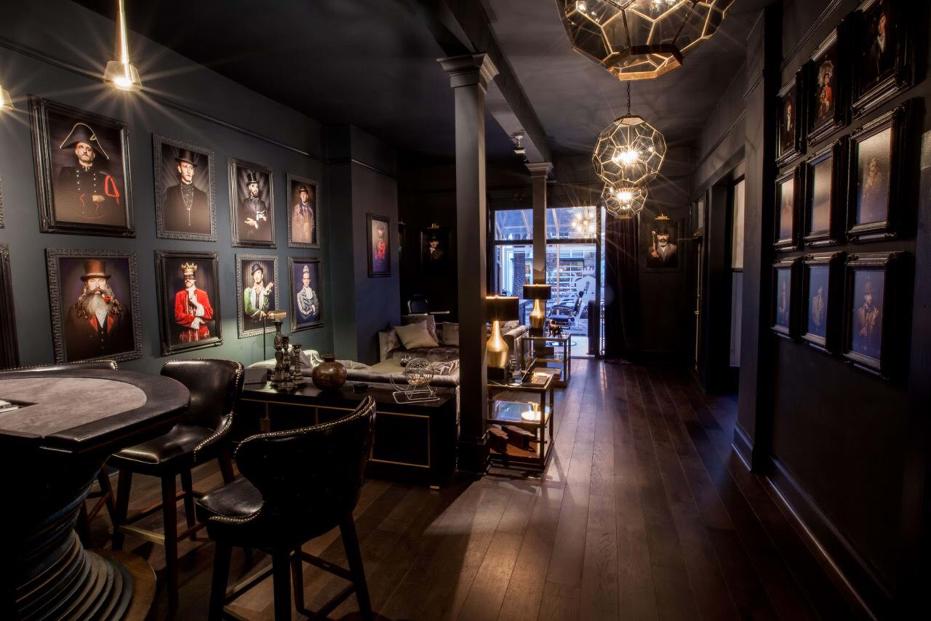 A dimly lit hallway with dark walls adorned with framed portraits, leading to a well-furnished seating area and a large window at the far end.